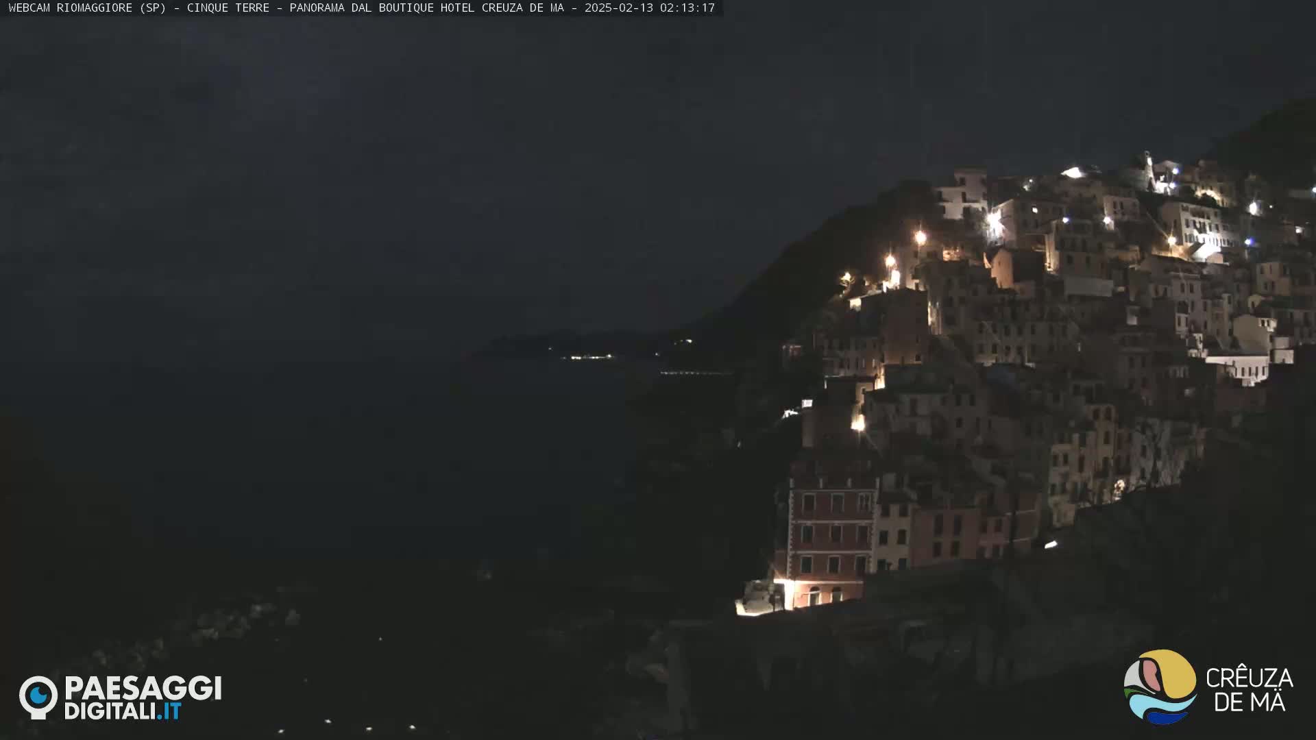 A nighttime view of a town built on a hillside overlooking a dark ocean under a clear, dark sky.