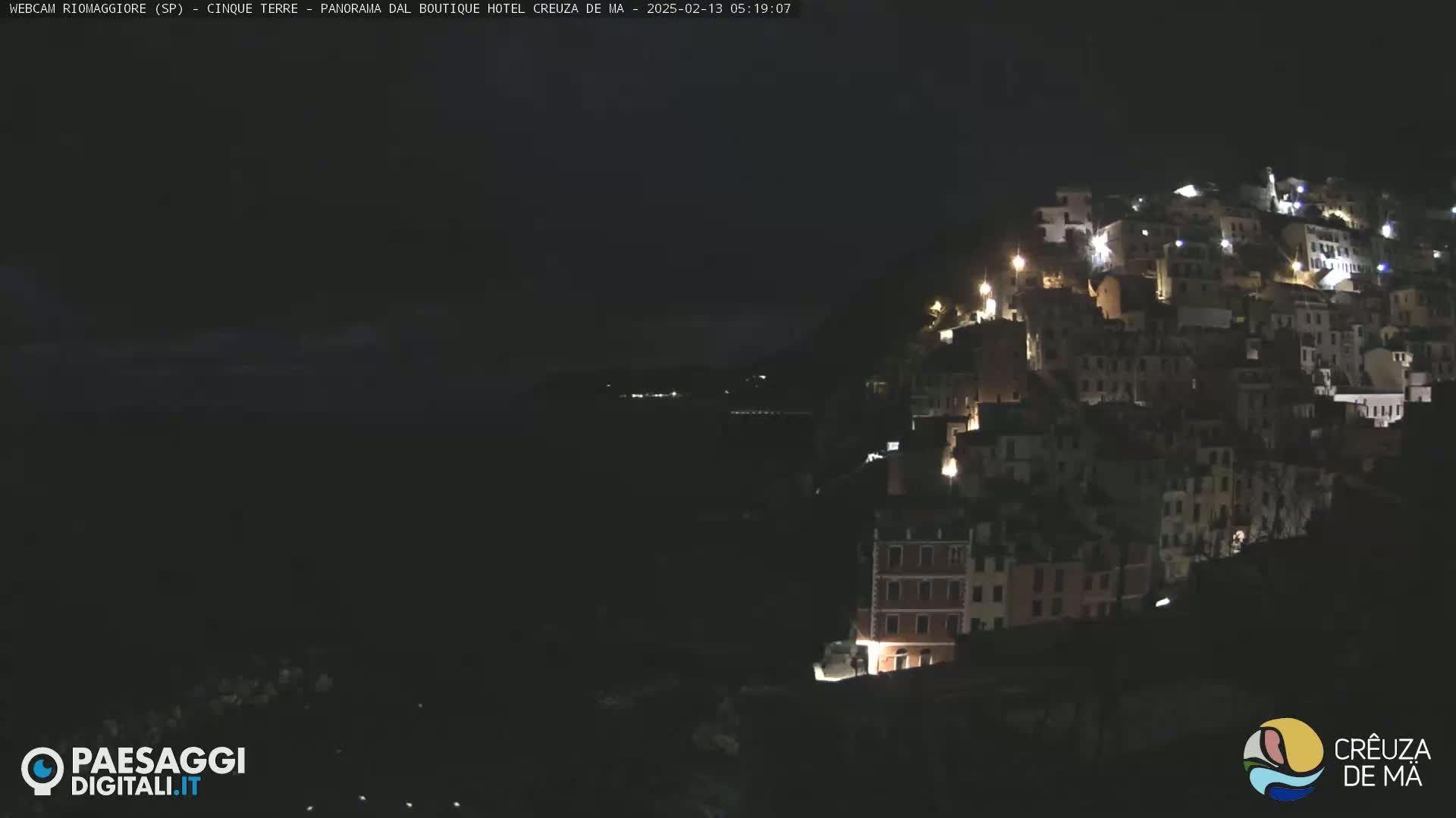 A nighttime view of a hillside village illuminated by lights, overlooking a dark body of water under a clear sky.