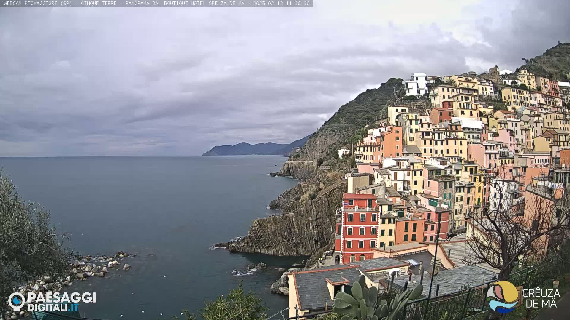 A coastal village of colorful buildings climbs a steep hillside overlooking a calm sea under a cloudy sky.