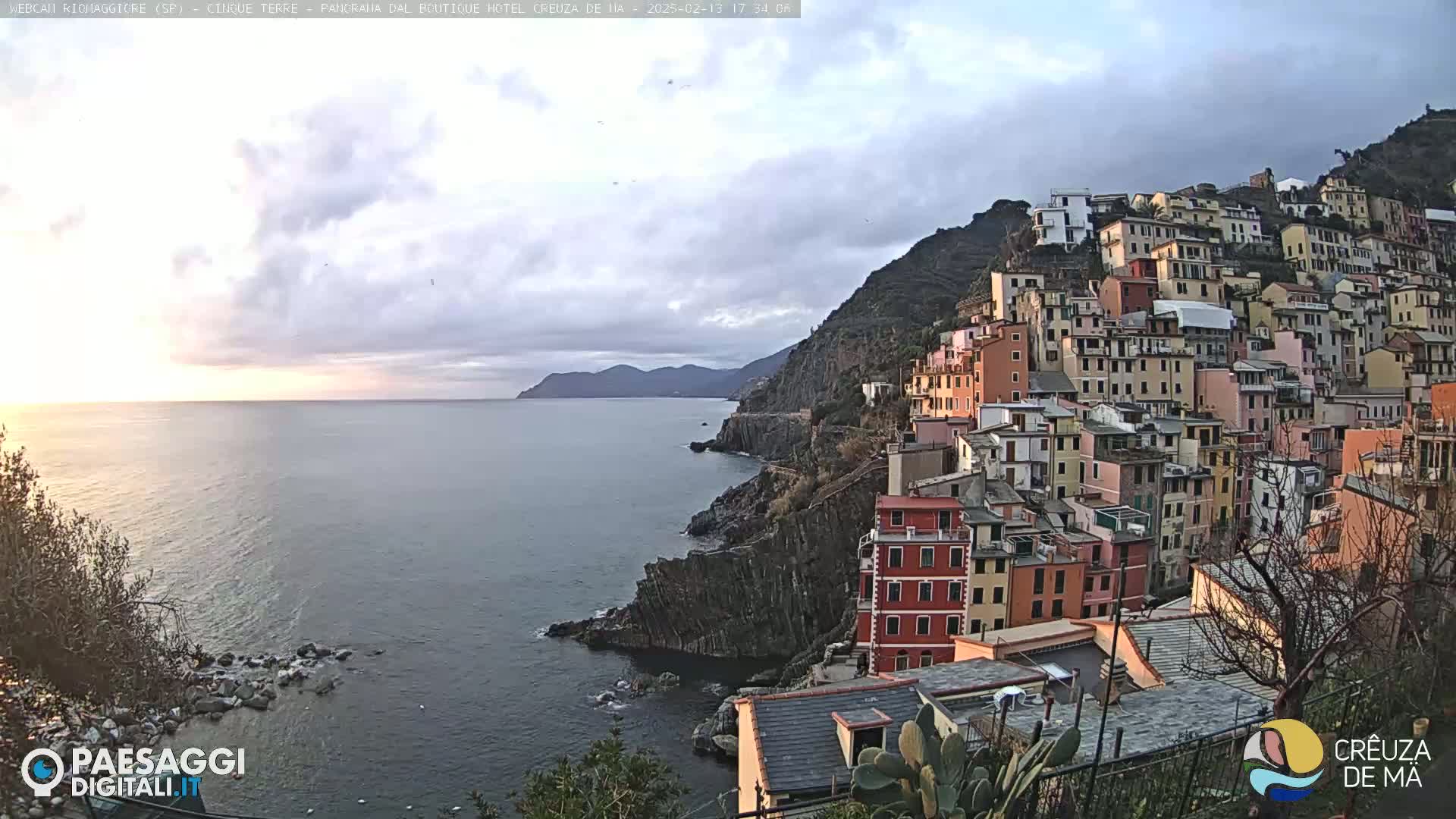 A coastal town of colorful buildings clings to a steep cliffside overlooking a calm sea under a mostly cloudy sky.