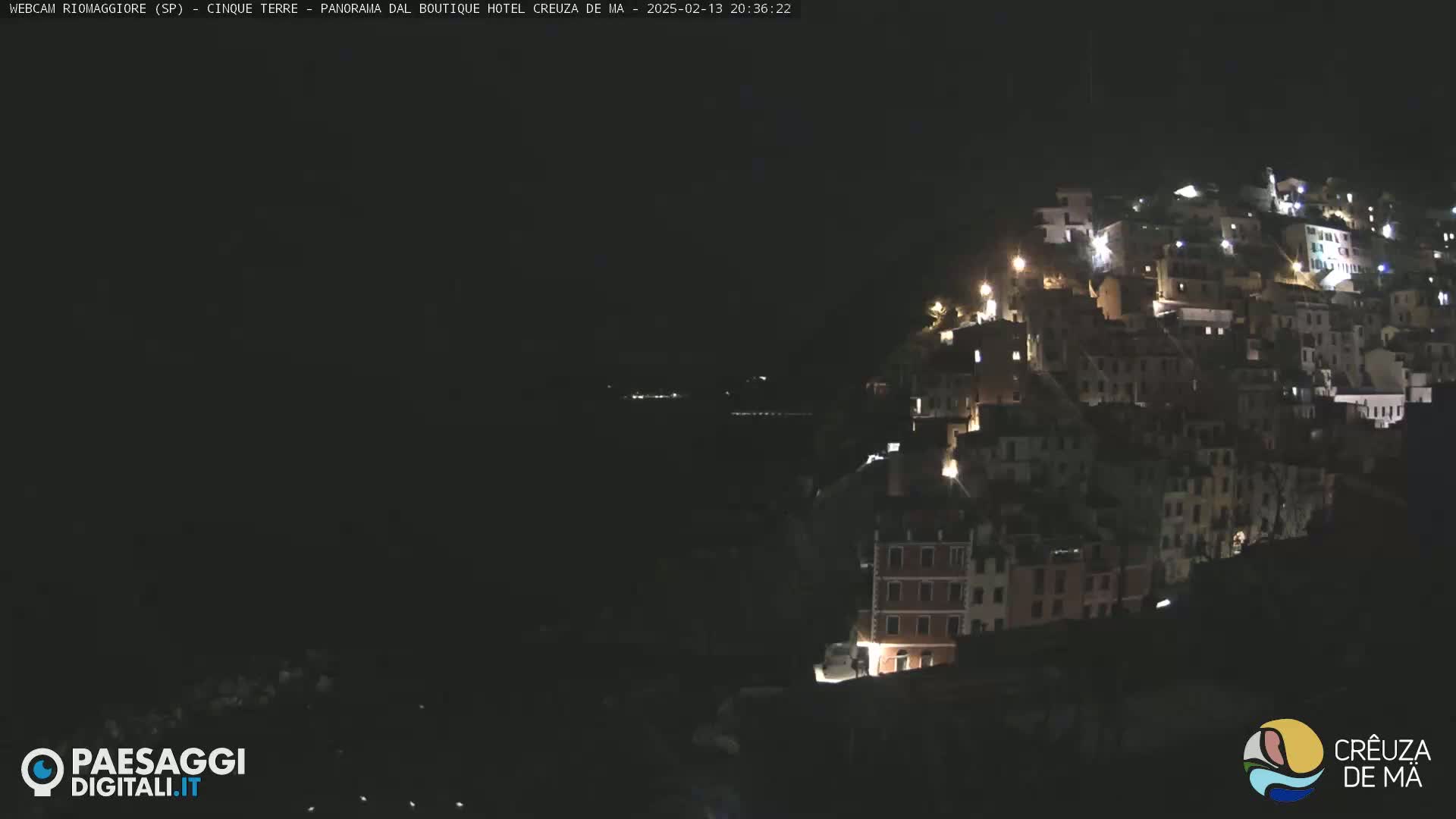 A nighttime view of a hillside town illuminated by lights, overlooking a dark body of water.