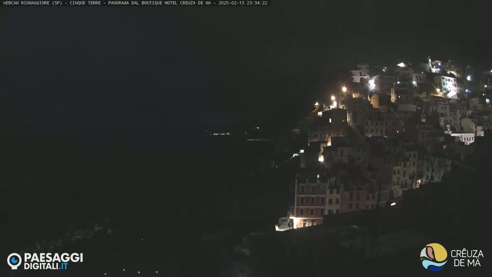 A nighttime view of a hillside town illuminated by lights, overlooking a dark body of water under a mostly clear, dark sky.