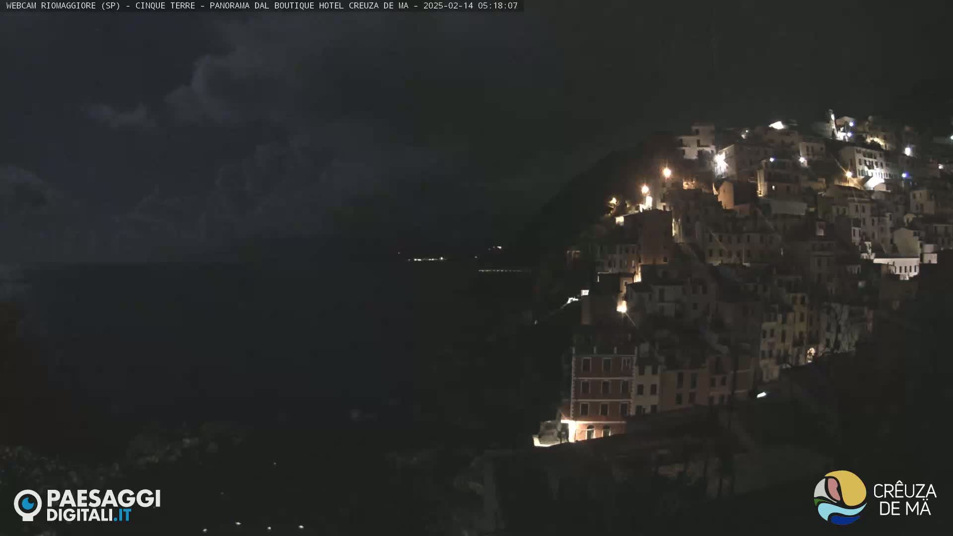 A nighttime view of a coastal town nestled on a hillside, illuminated by lights, under a partly cloudy sky.