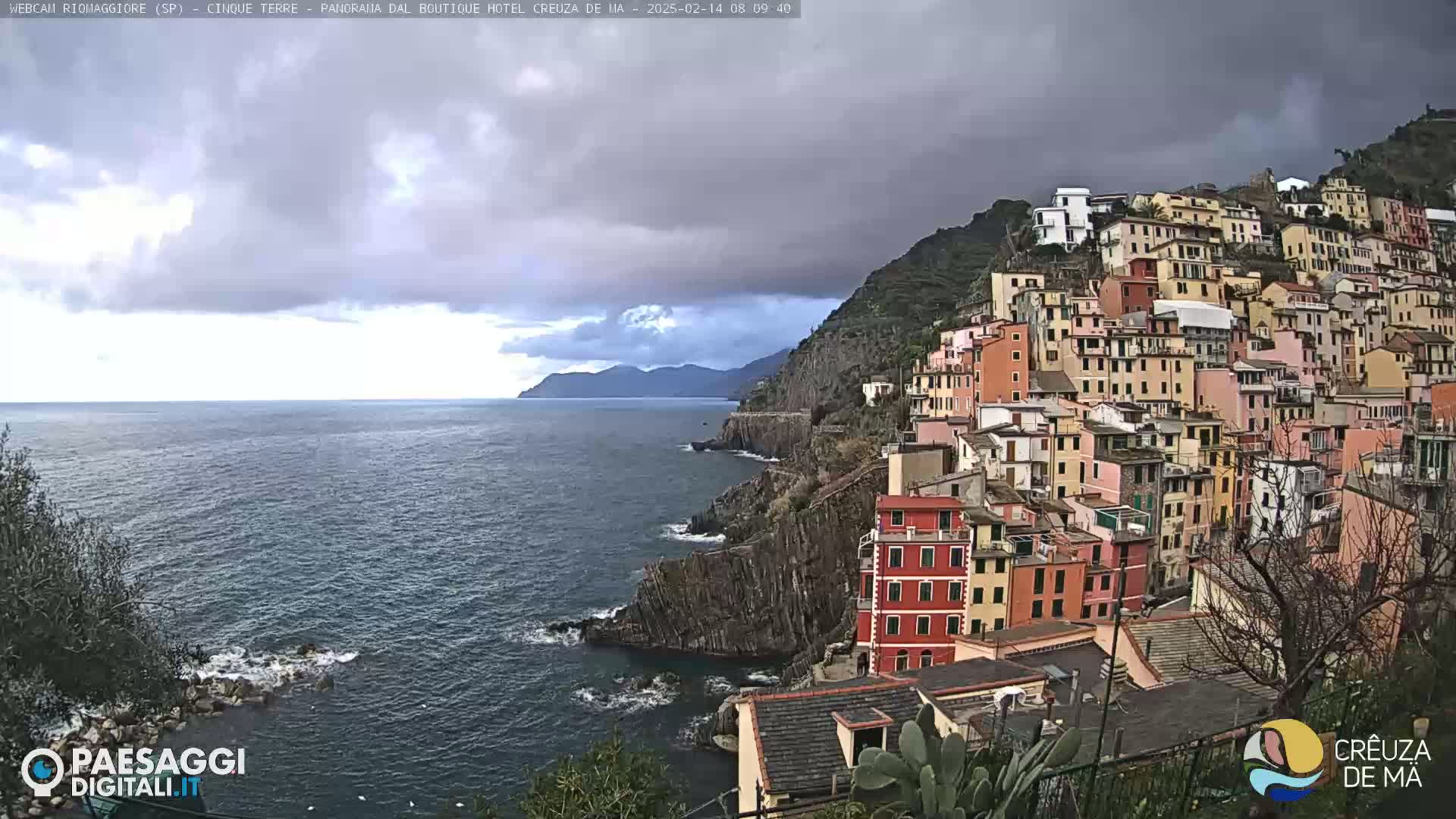 A colorful village clings to a steep, rocky cliff overlooking a dark blue sea under an overcast sky.