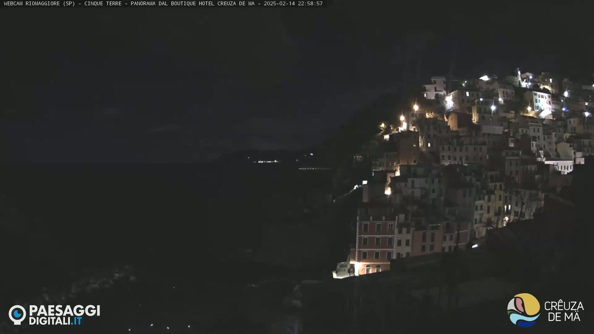 A nighttime, coastal town with many illuminated buildings is nestled on a hillside overlooking a dark body of water.