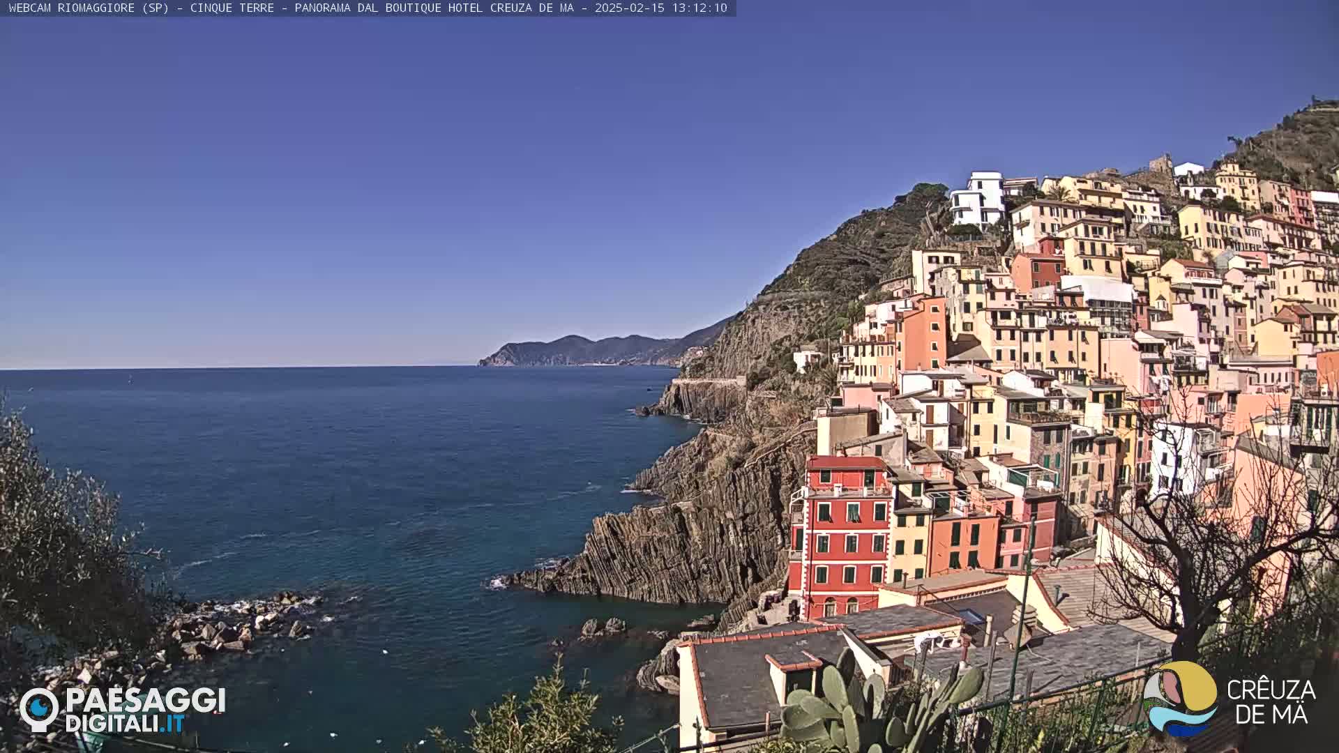 A colorful hillside village clings to a rocky coastline overlooking a calm, blue sea under a clear sky.