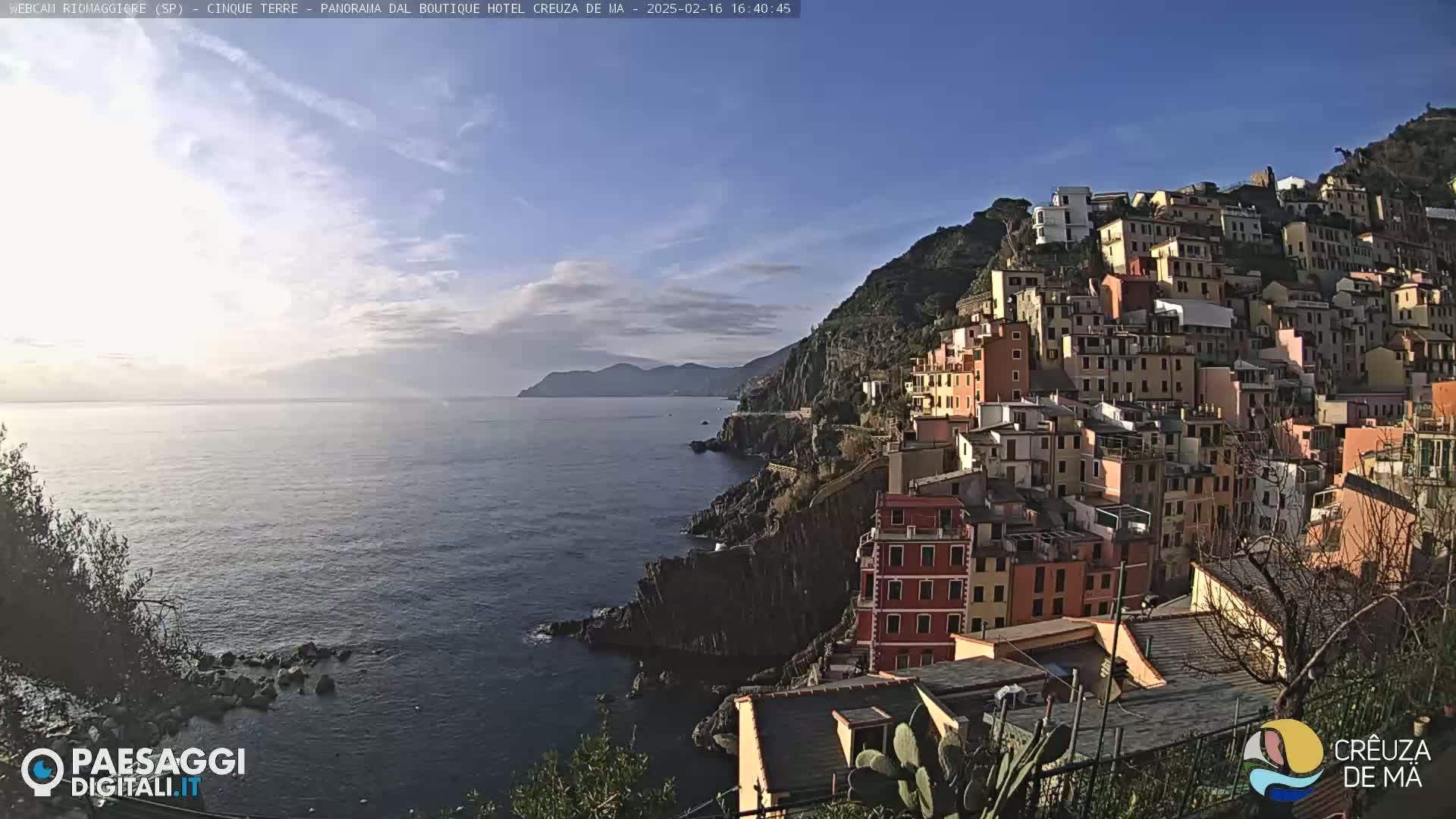A coastal town with colorful buildings clinging to a steep hillside overlooks a calm sea under a clear, sunny sky.