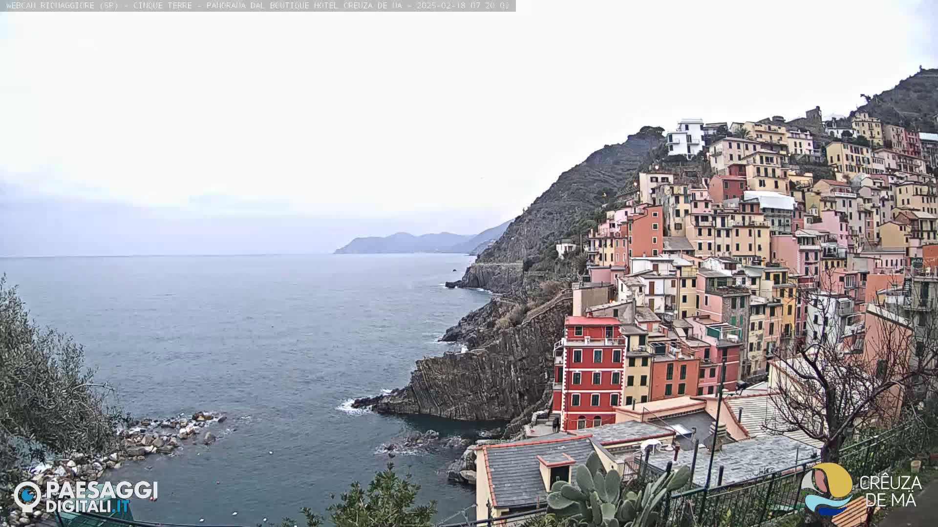 A colorful hillside town clings to a steep cliff overlooking a calm, gray sea under an overcast sky.