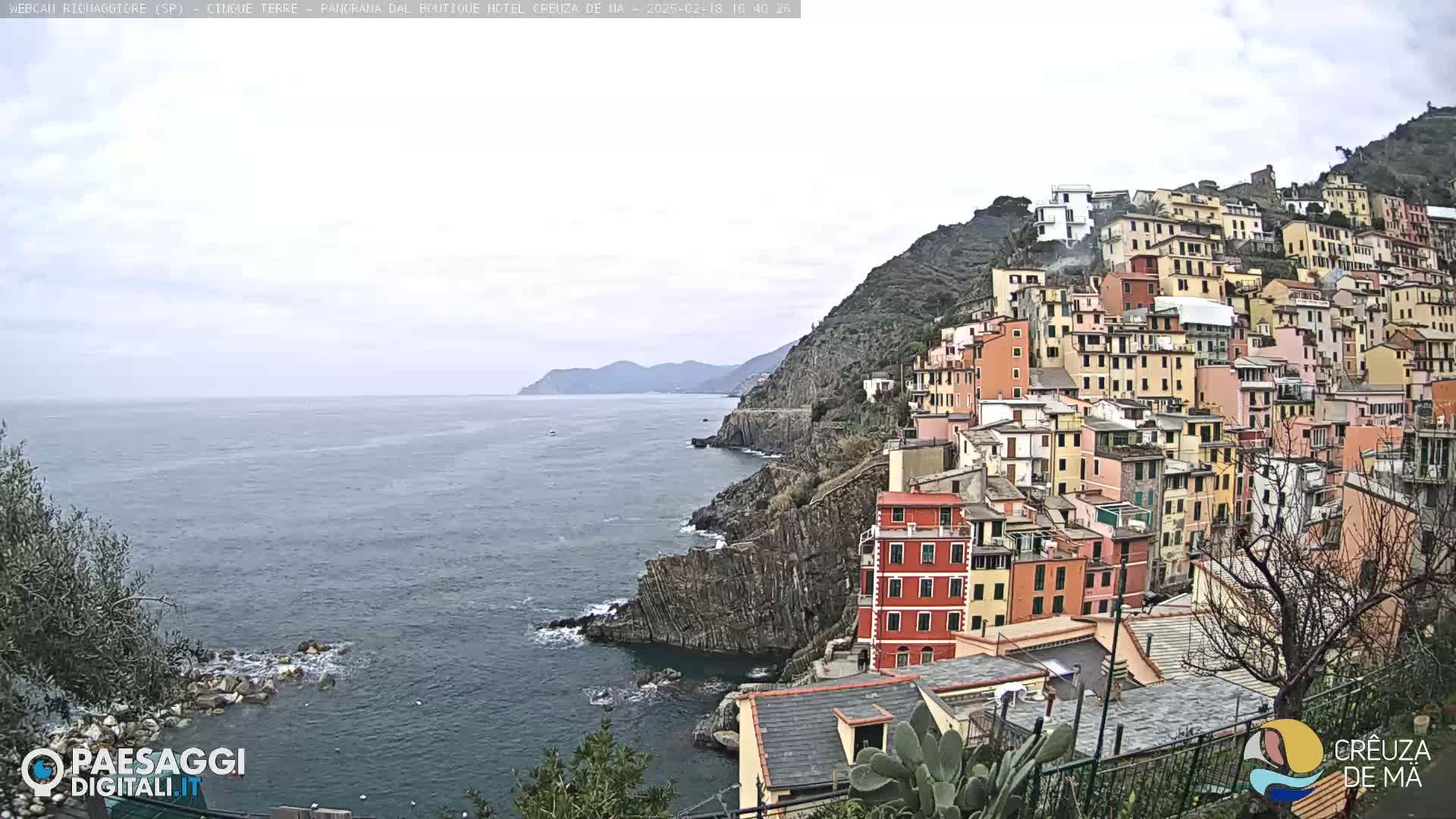 A coastal town of colorful buildings clings to a steep hillside overlooking a calm sea under a cloudy sky.