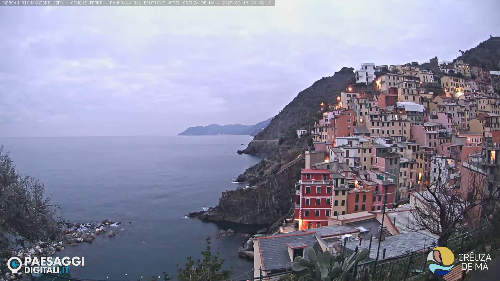 A coastal village of colorful buildings clinging to a steep hillside overlooks a calm sea under a cloudy twilight sky.