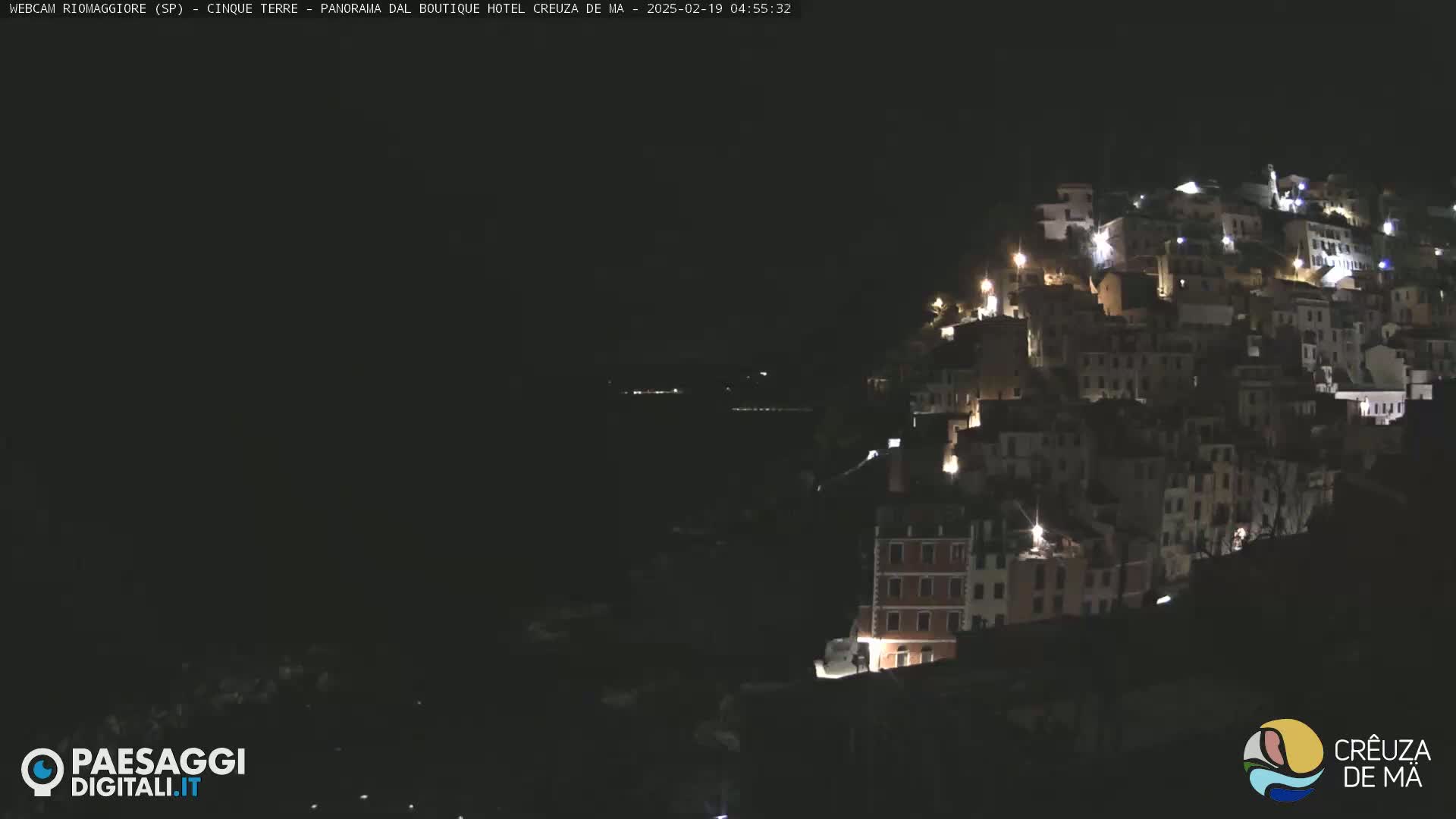 A nighttime view of a hillside town illuminated by lights, overlooking a dark body of water under a clear, dark sky.