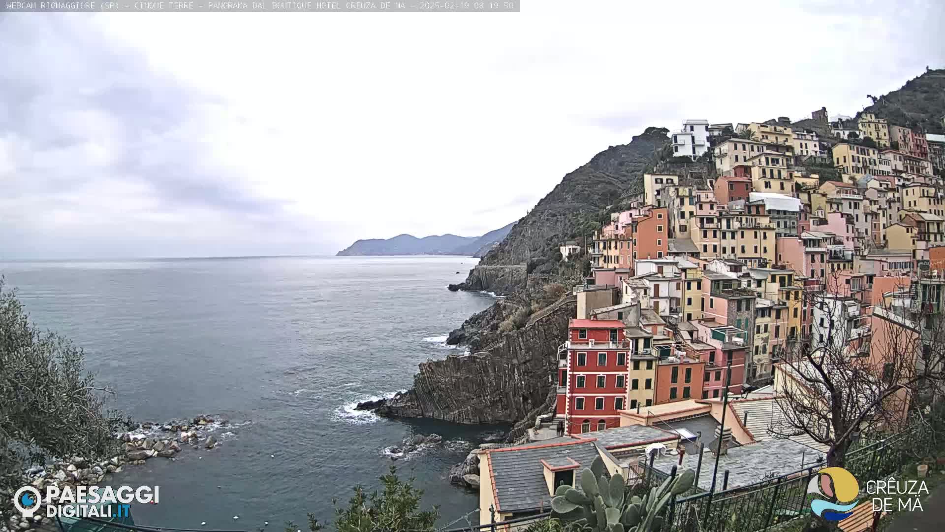 A coastal town of colorful buildings clinging to a steep hillside overlooks a calm sea under an overcast sky.