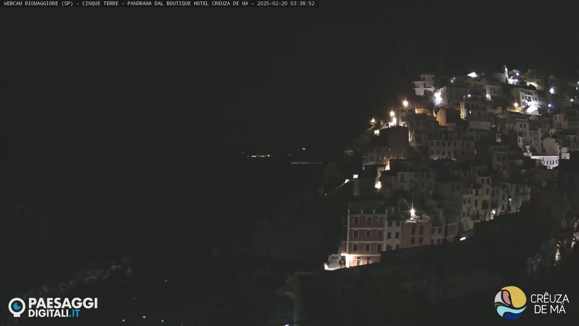 A nighttime view of a hillside town illuminated by lights, overlooking a dark body of water under a clear, dark sky.