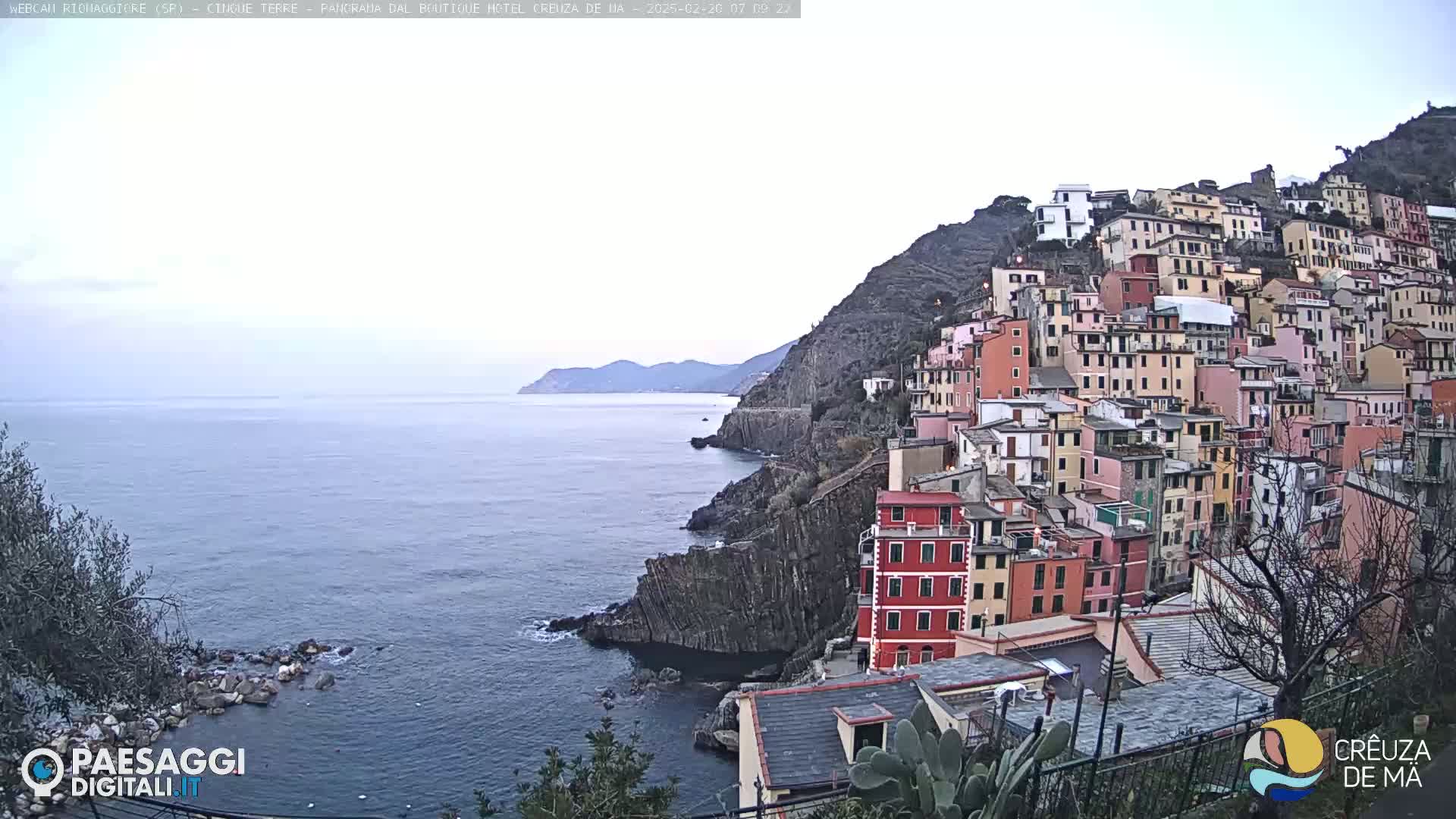 A coastal town with colorful buildings clinging to a steep hillside overlooks a calm sea under an overcast sky.