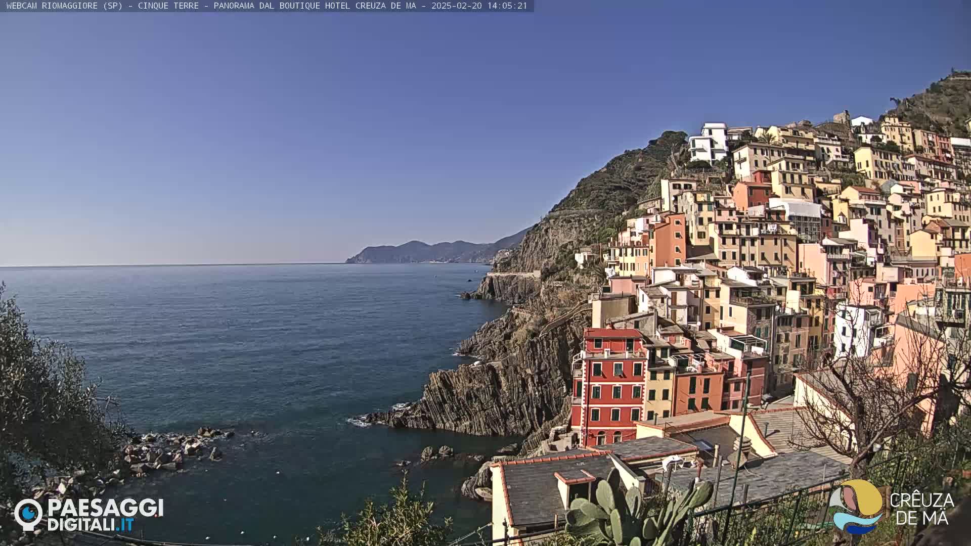 A colorful village clings to a steep, rocky hillside overlooking a calm blue sea under a clear blue sky.