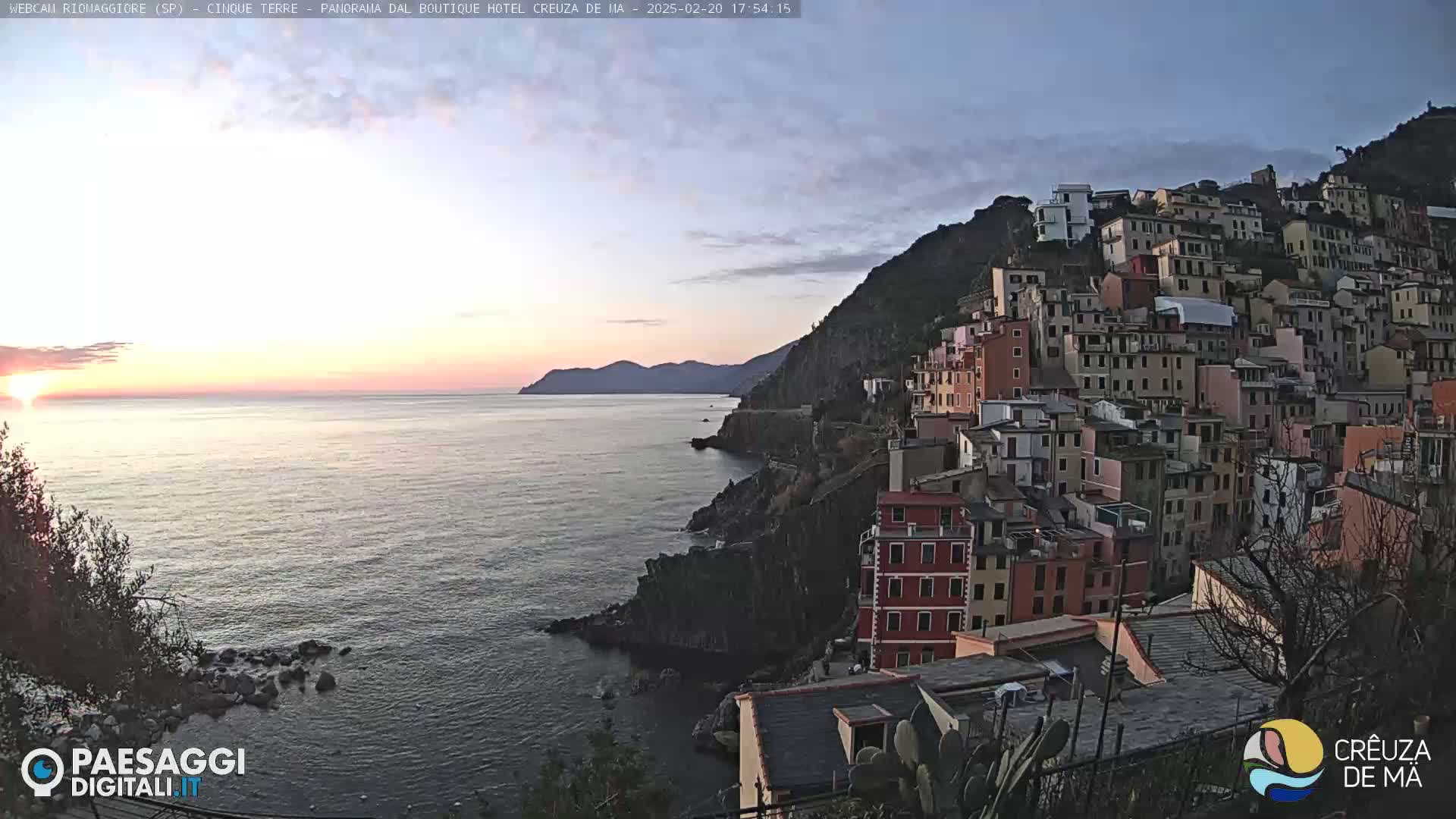 A coastal town built on a steep hillside overlooks a calm sea at sunset.