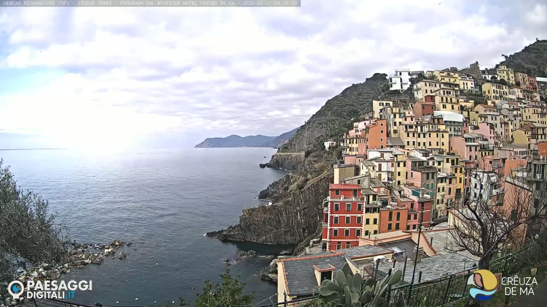 A coastal town with colorful buildings stacked on a hillside overlooks a calm sea under a cloudy sky.