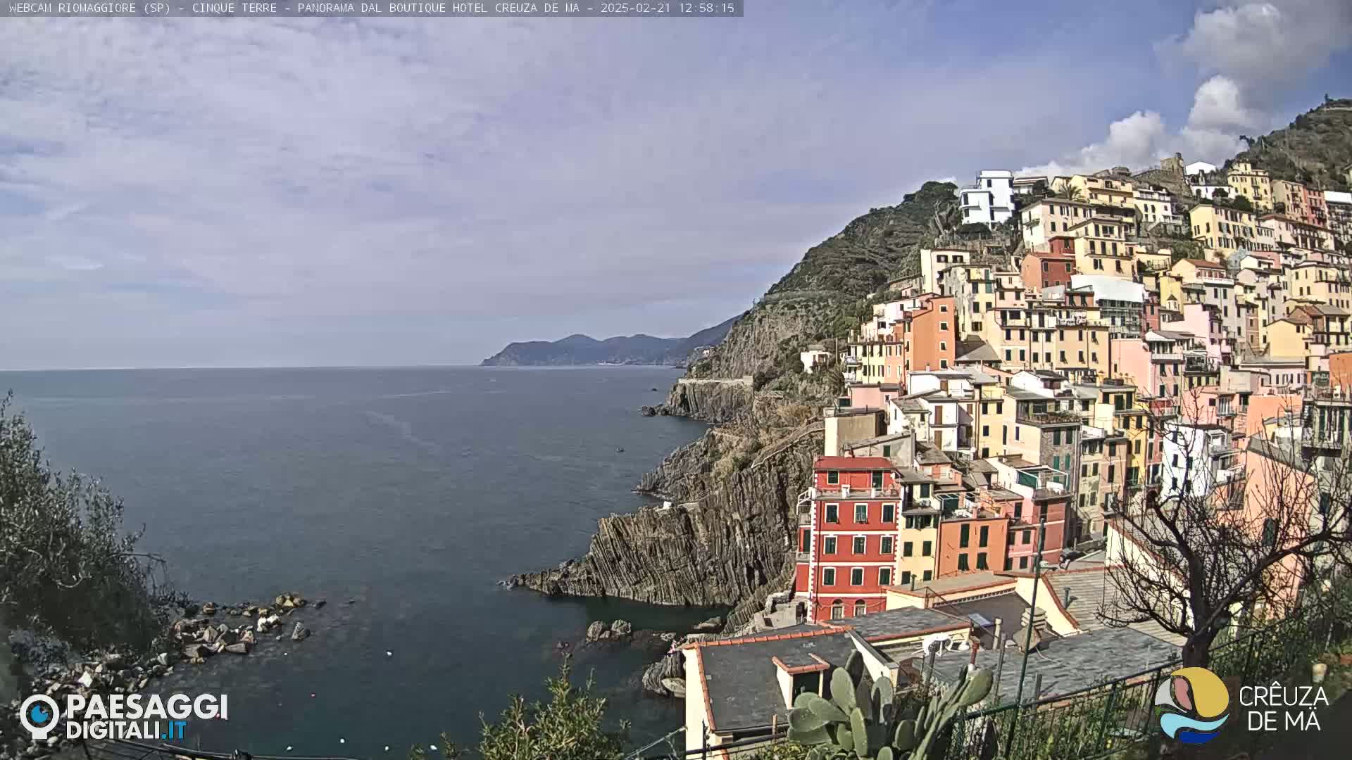 A coastal town with colorful buildings clinging to a steep hillside overlooks a calm sea under a partly cloudy sky.