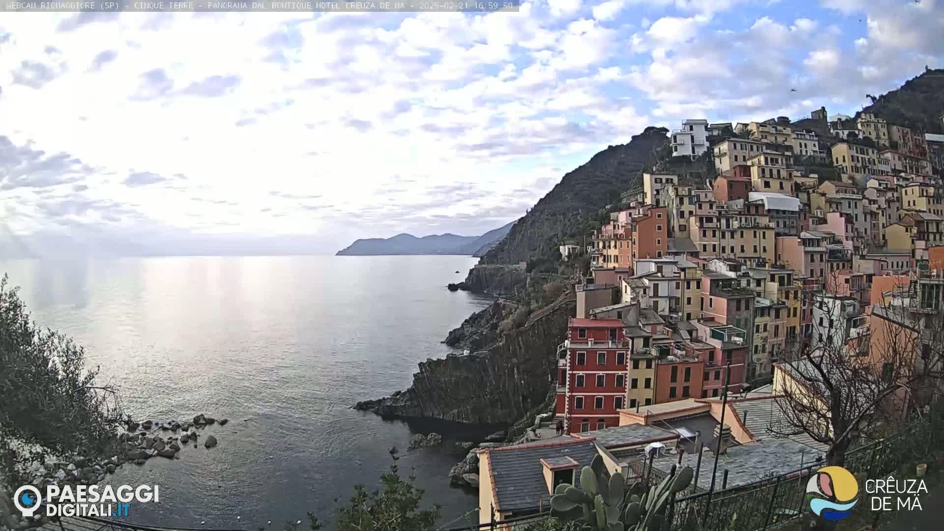 A picturesque coastal town, clinging to a steep hillside, overlooks a calm sea under a partly cloudy sky.