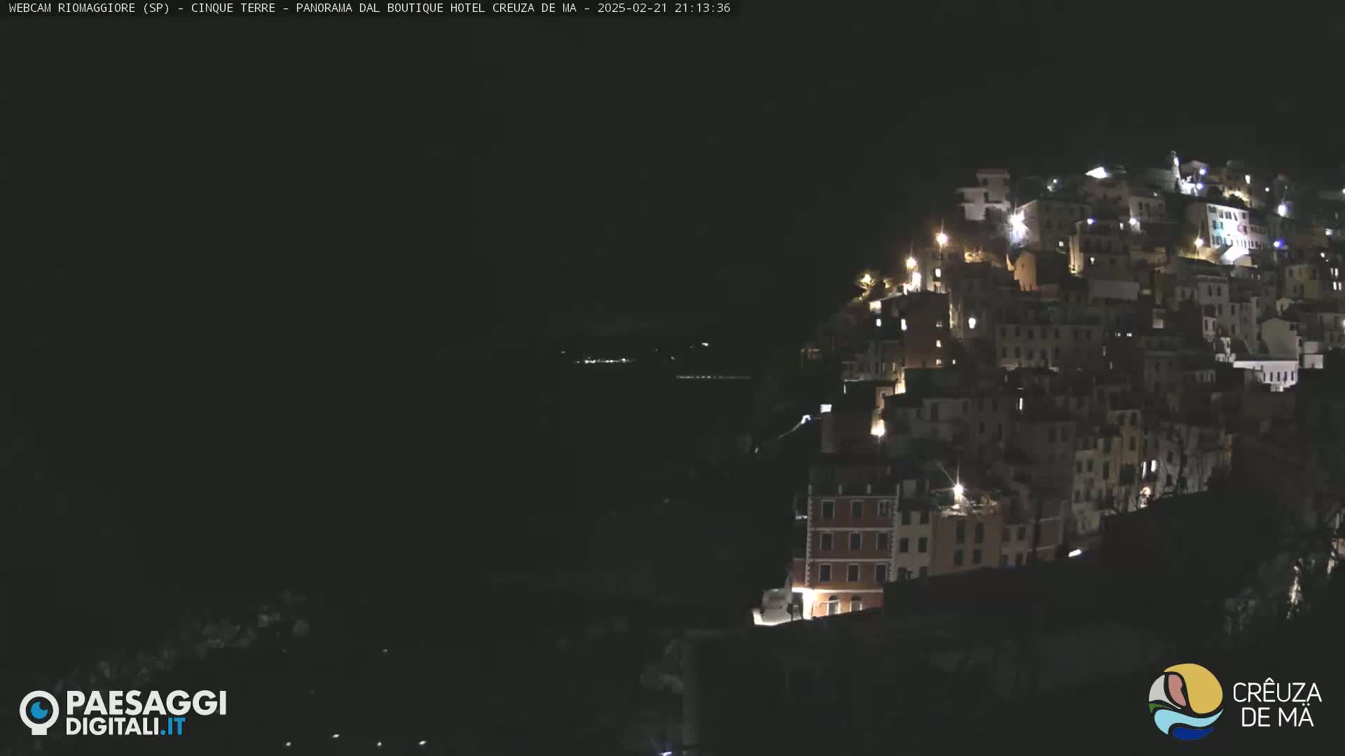 A nighttime view of a hillside town illuminated by lights, overlooking a dark body of water.