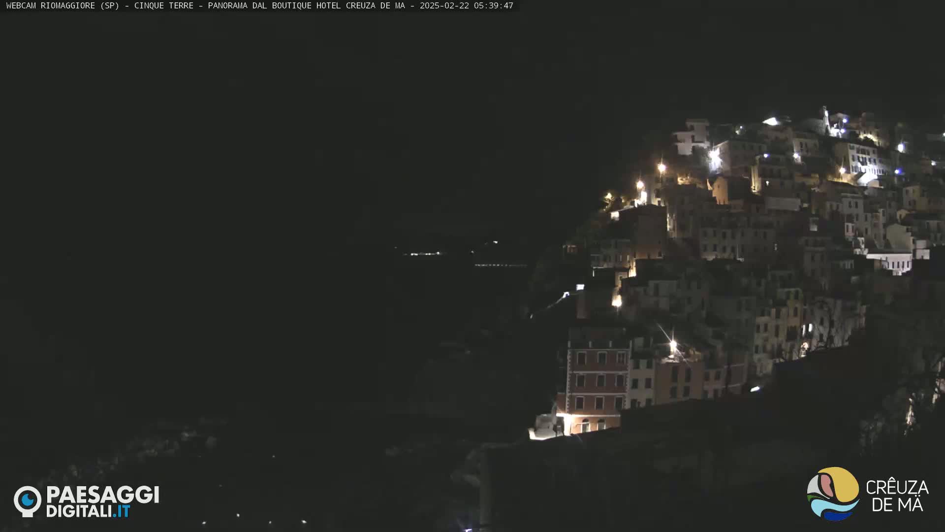 A nighttime view of a hillside town illuminated by lights, overlooking a dark body of water.