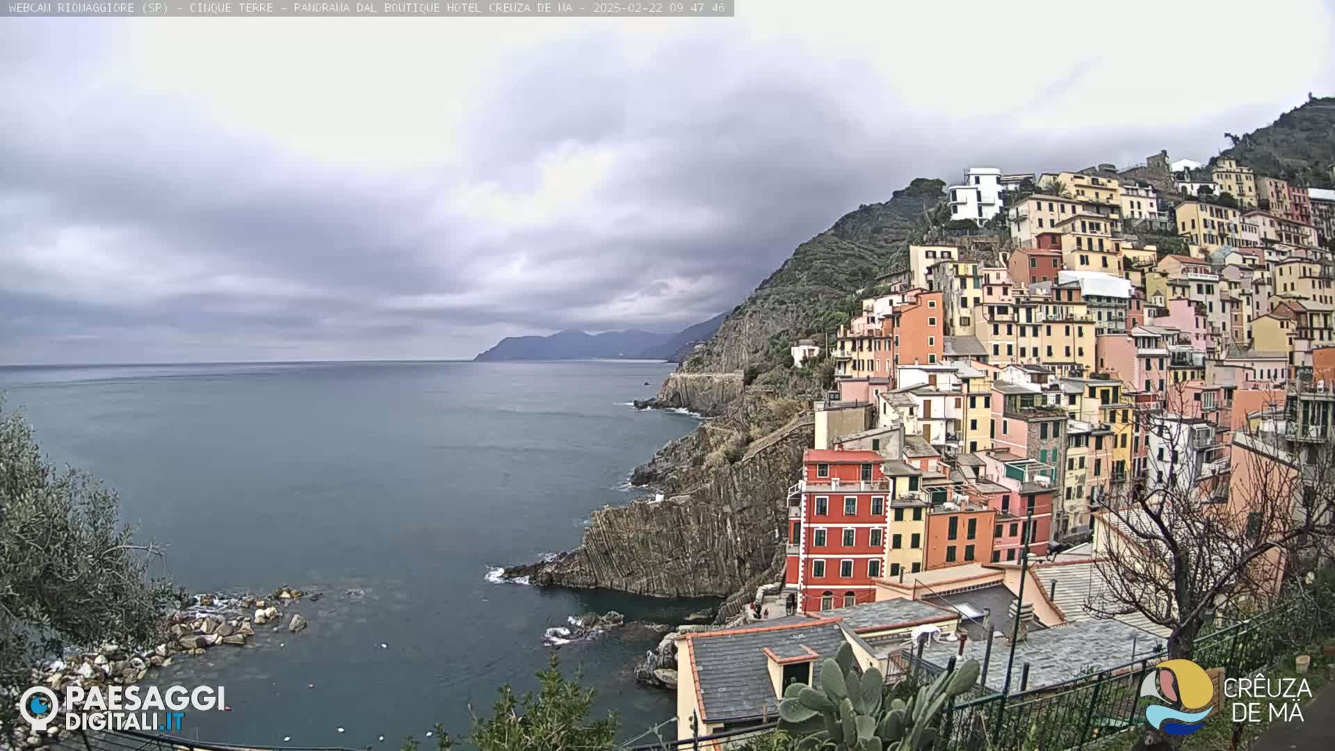 A coastal village of colorful buildings clings to a steep hillside overlooking a calm sea under a cloudy sky.
