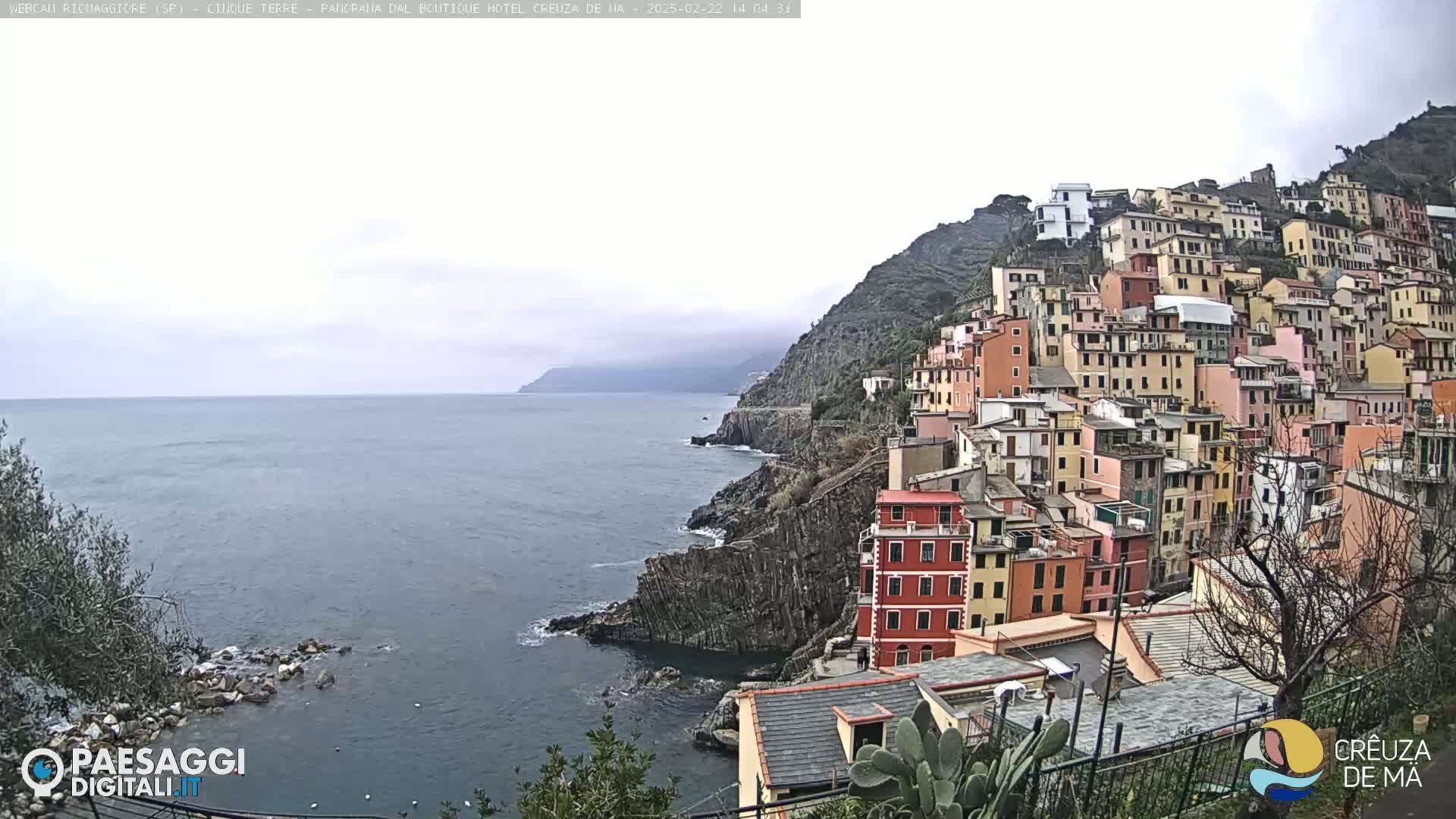 A coastal village with colorful buildings stacked on a steep hillside overlooks a calm sea under an overcast sky.