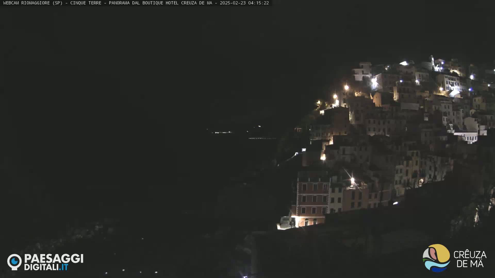 A nighttime view of a hillside town illuminated by lights, overlooking a dark body of water.