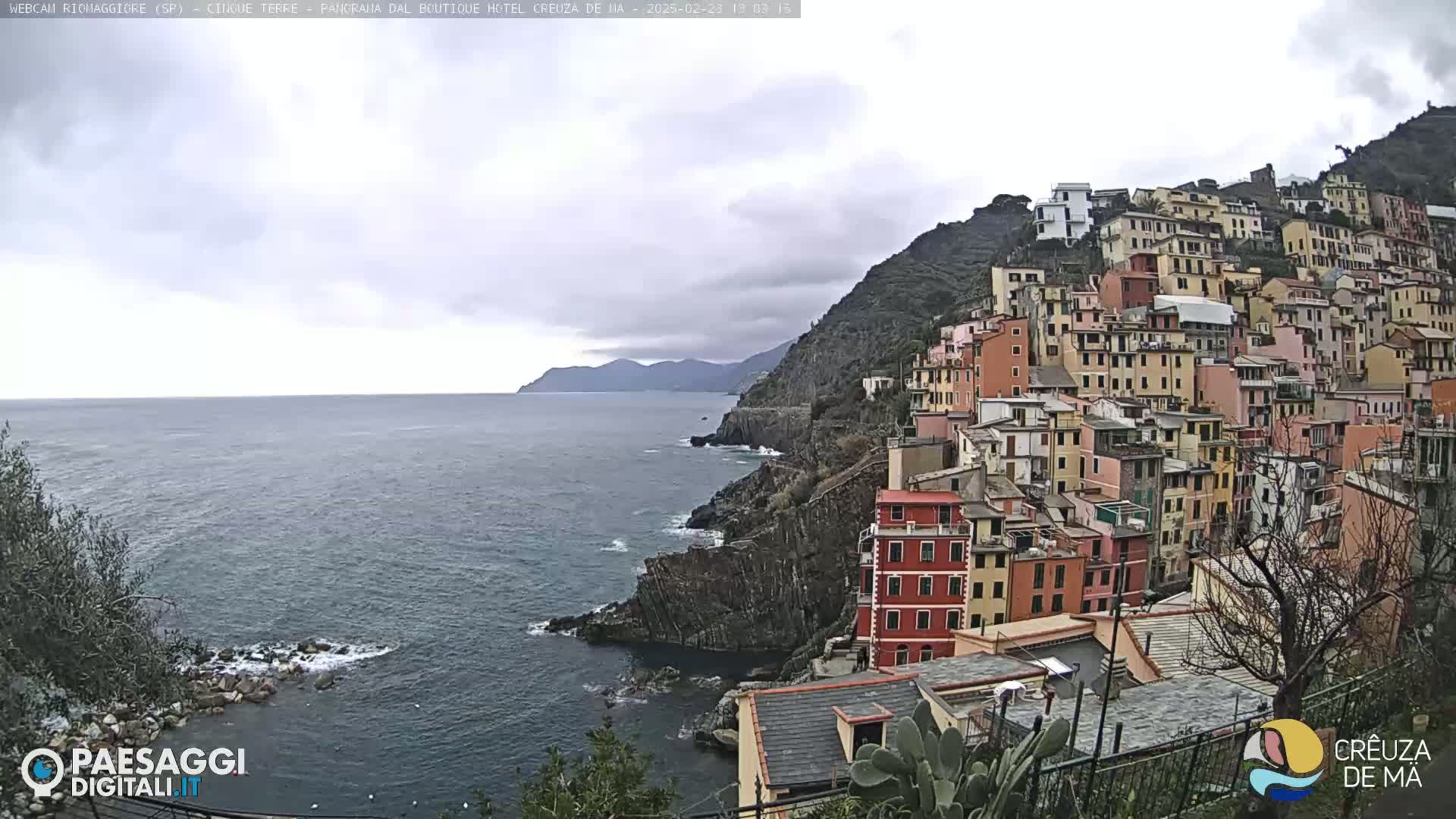 A coastal village of colorful buildings clings to a steep hillside overlooking a calm, overcast sea.