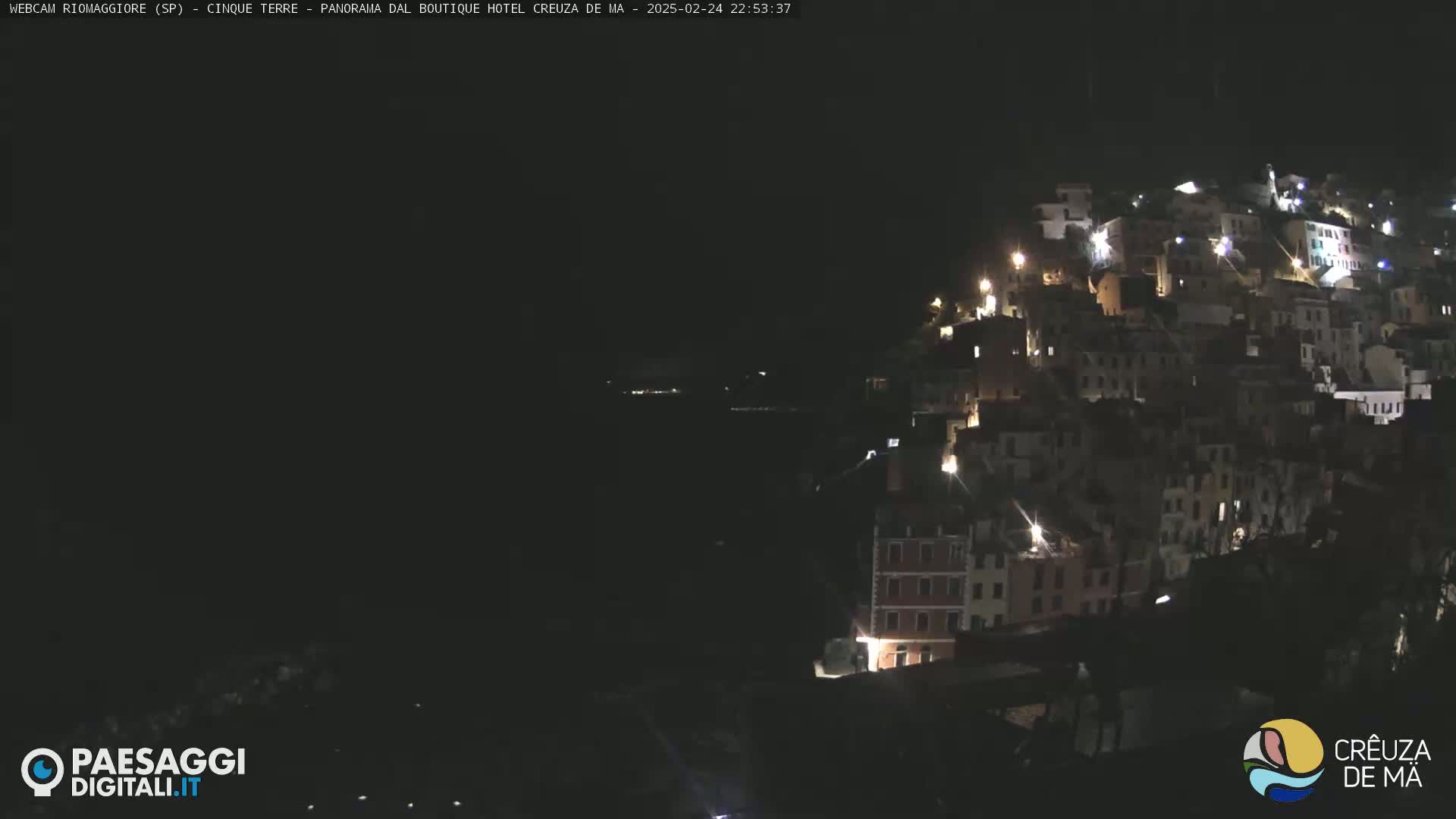 A nighttime view of a hillside town, illuminated by scattered lights, overlooking a dark body of water.