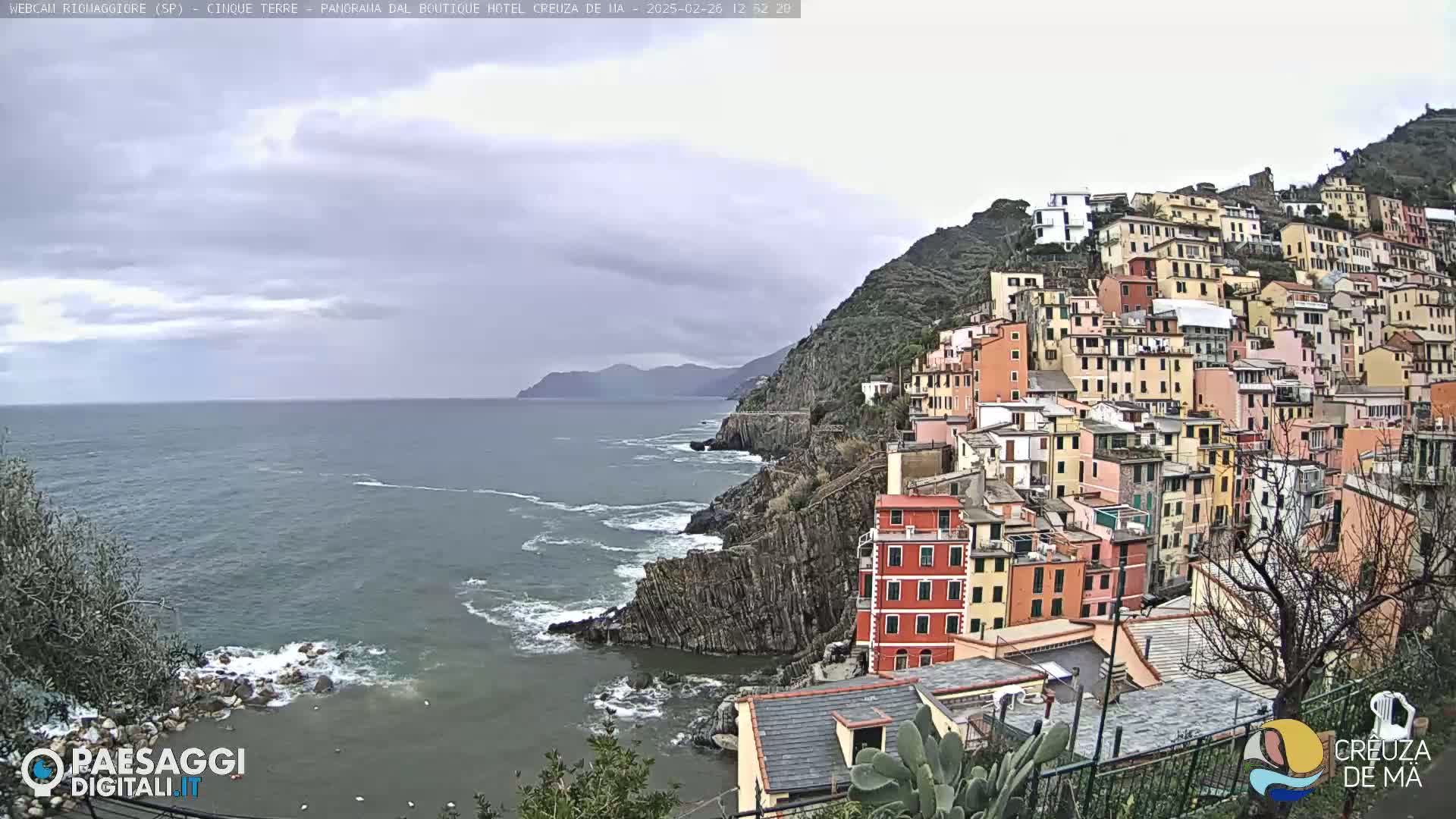 A colorful village clings to a steep, rocky hillside overlooking a gray, choppy sea under an overcast sky.