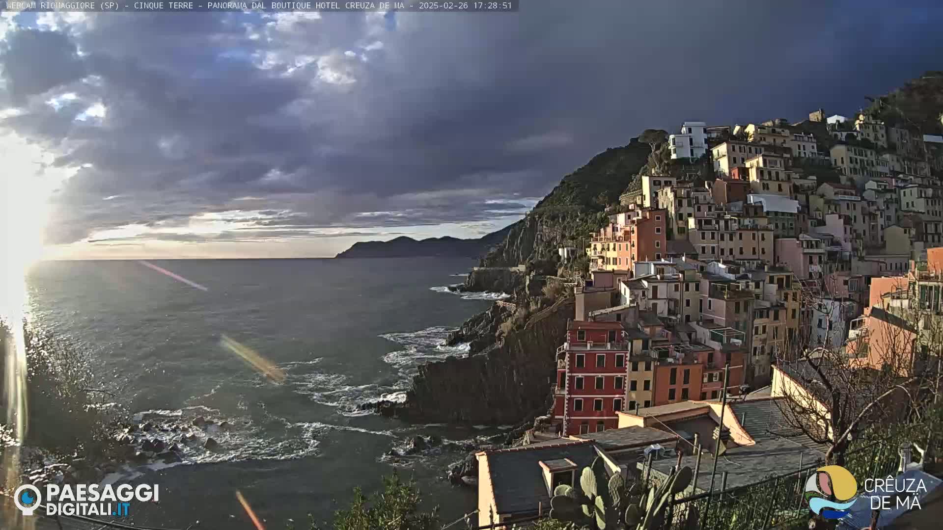 A coastal town built on a steep hillside overlooks a dark, wavy sea under a partly cloudy sky.