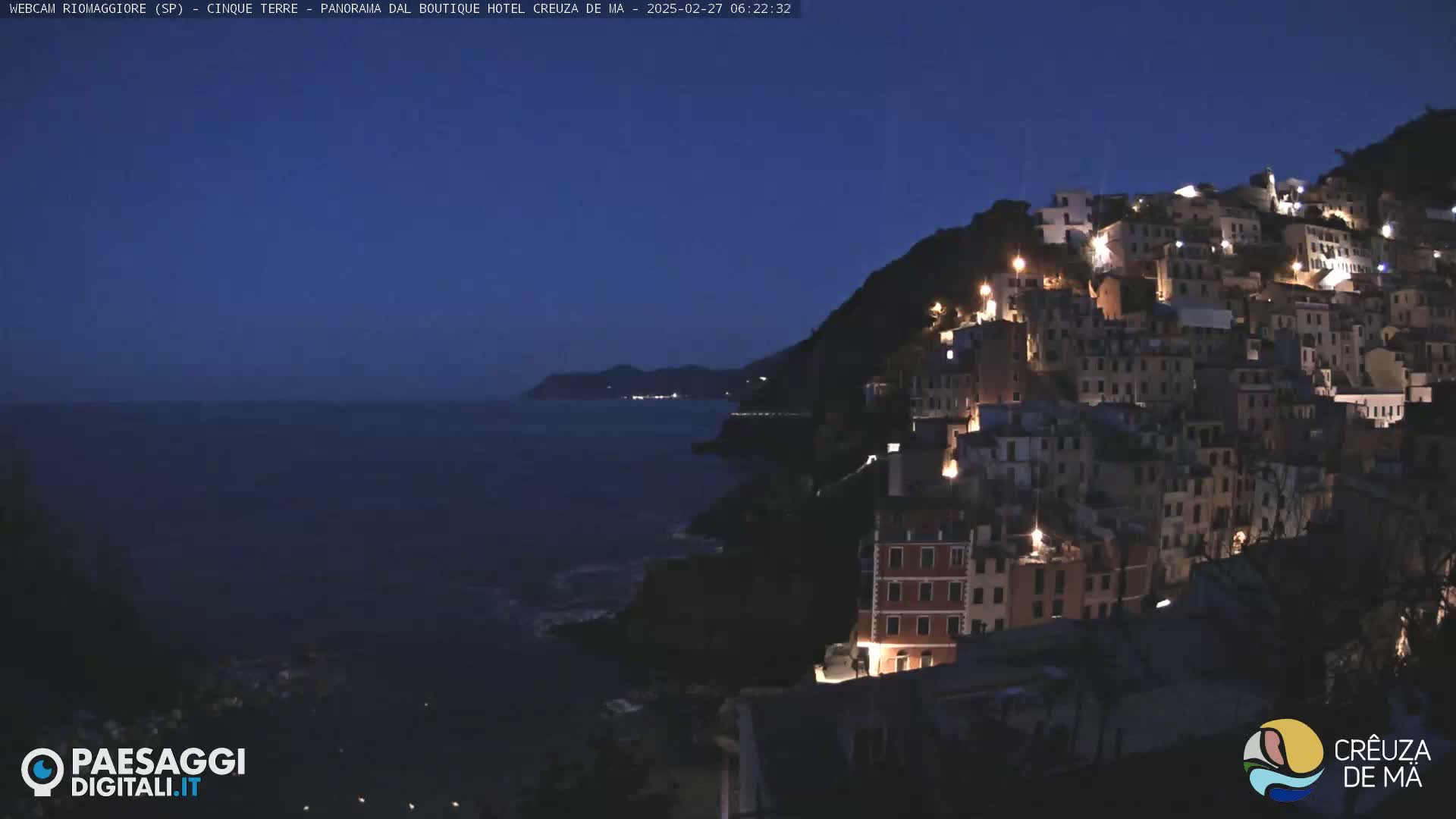 A nighttime coastal village clings to a hillside overlooking a dark sea under a twilight sky.