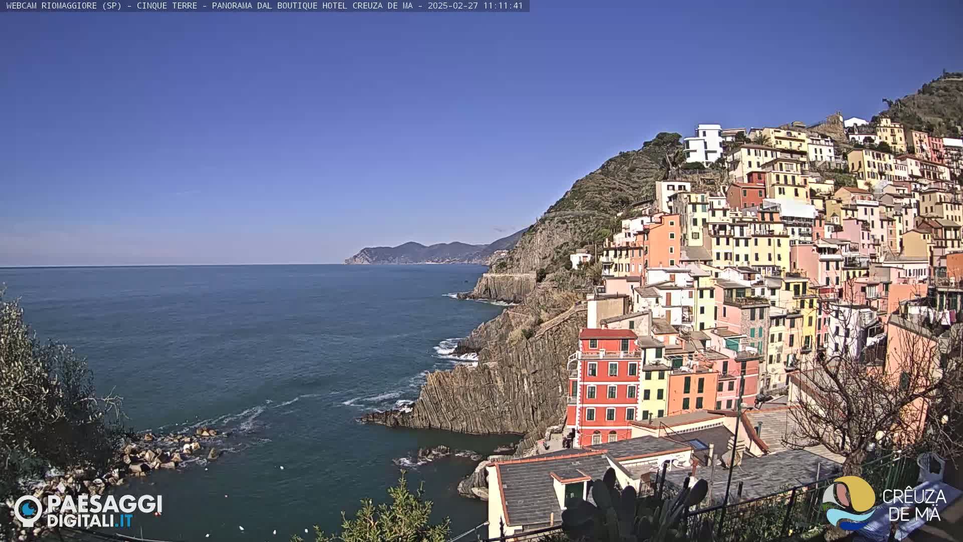 A colorful village clings to a steep hillside overlooking a calm blue sea under a clear blue sky.