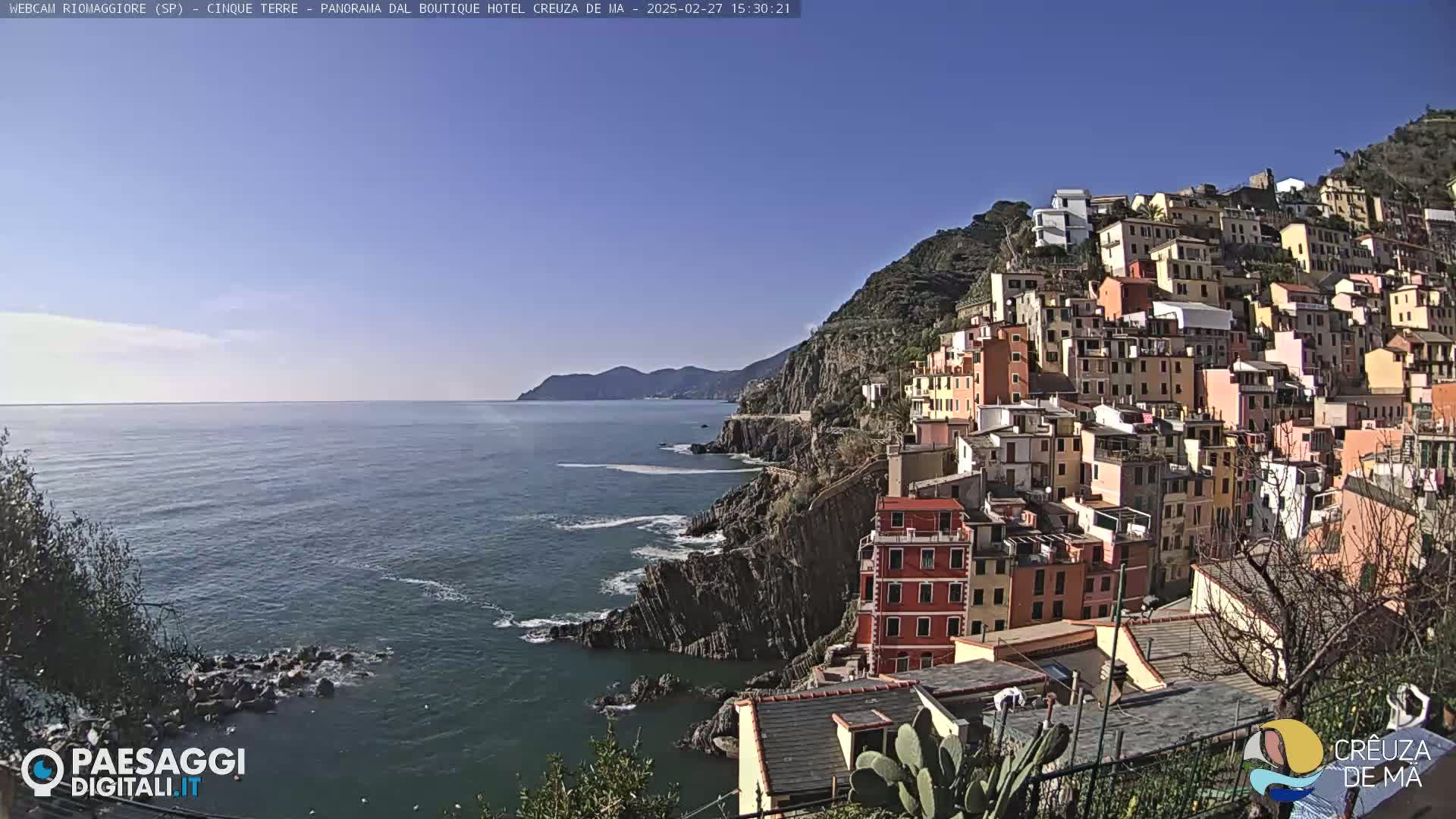 A colorful village clings to a steep cliffside overlooking a calm blue sea under a clear, sunny sky.