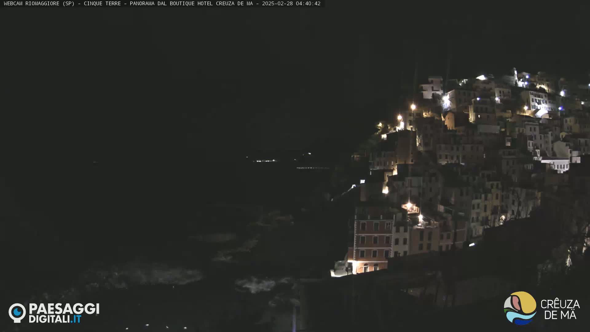 A nighttime view of a hillside town illuminated by lights, overlooking a dark body of water.
