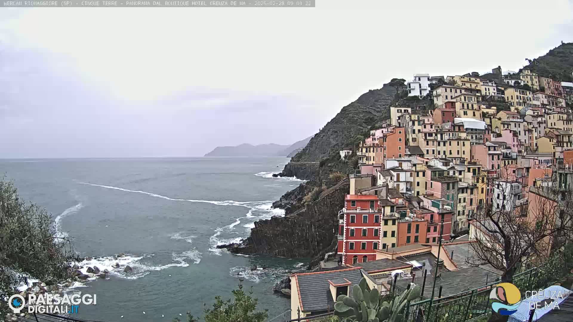 A coastal town of colorful buildings clings to a steep hillside overlooking a choppy gray sea under an overcast sky.
