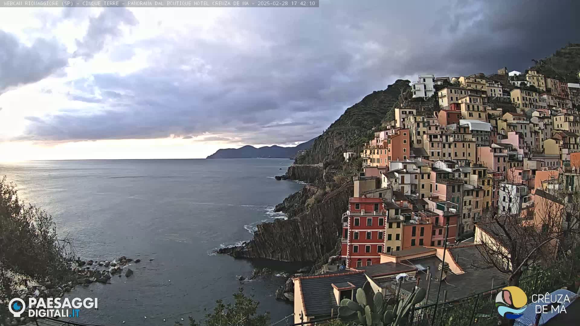 A coastal village of colorful buildings climbs a steep hillside overlooking a calm sea under a cloudy sky.