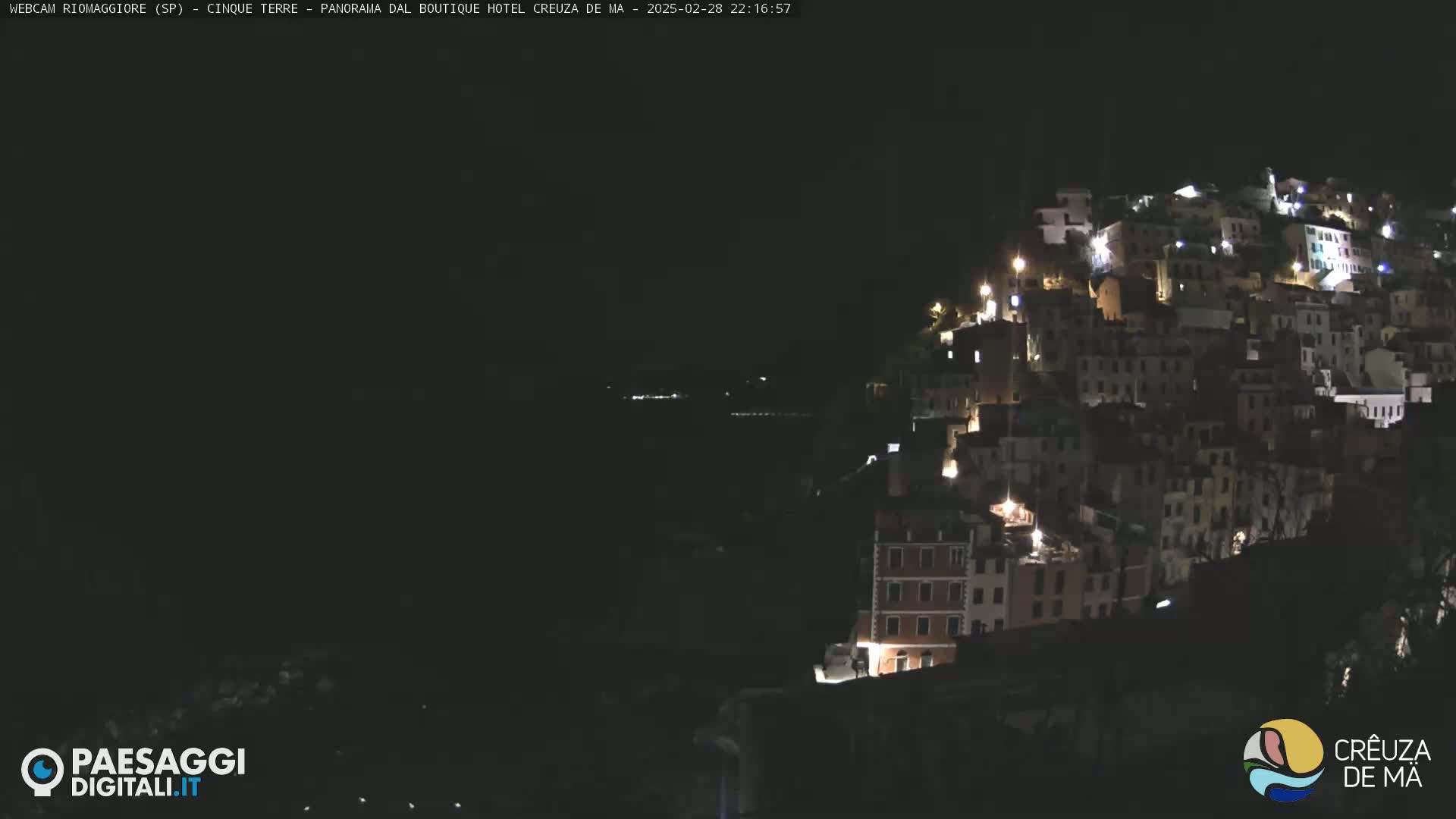 A nighttime view of a hillside town illuminated by lights, with a dark body of water visible in the distance.