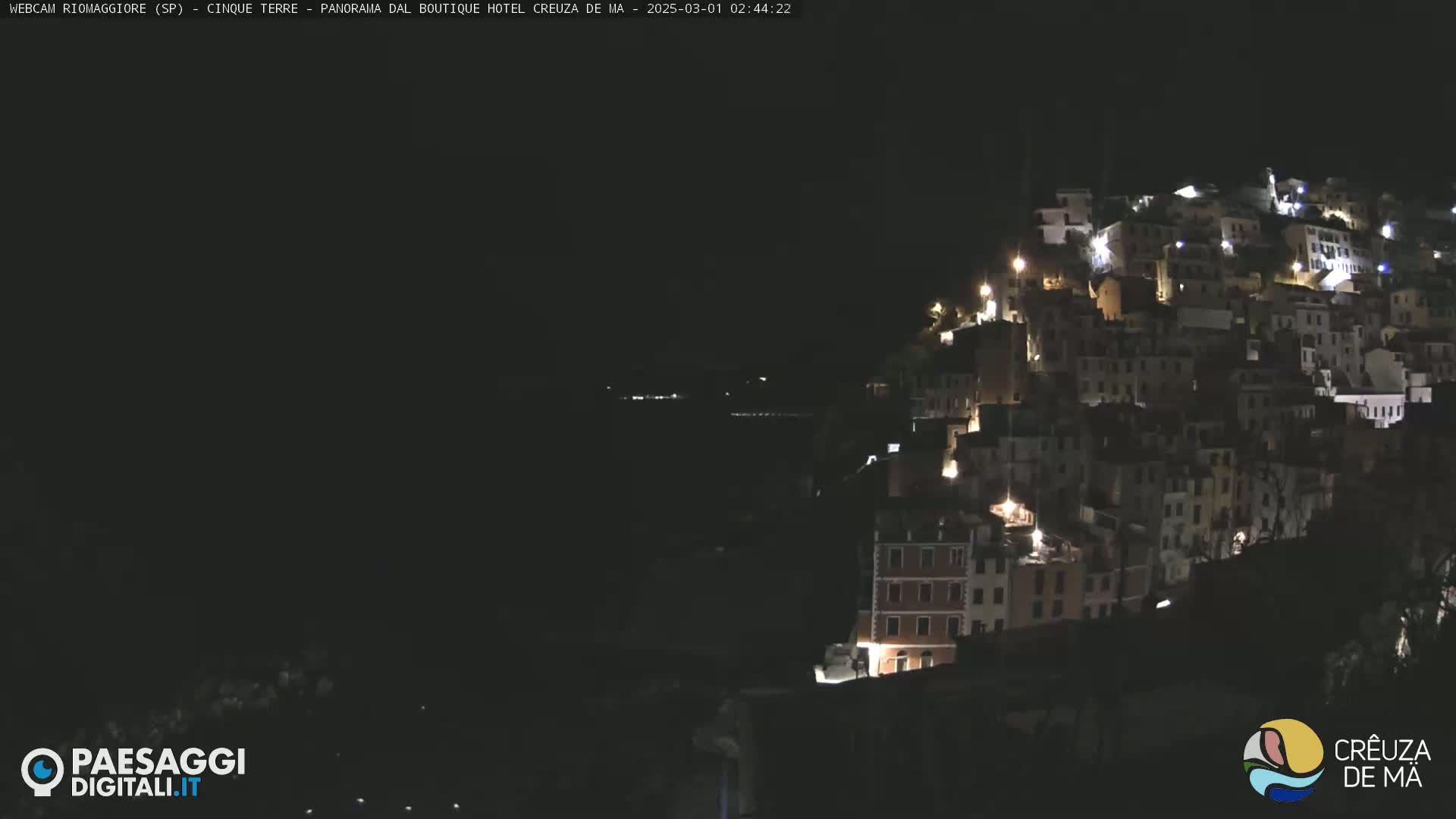 A hillside town is illuminated at night, with the dark ocean visible in the foreground.