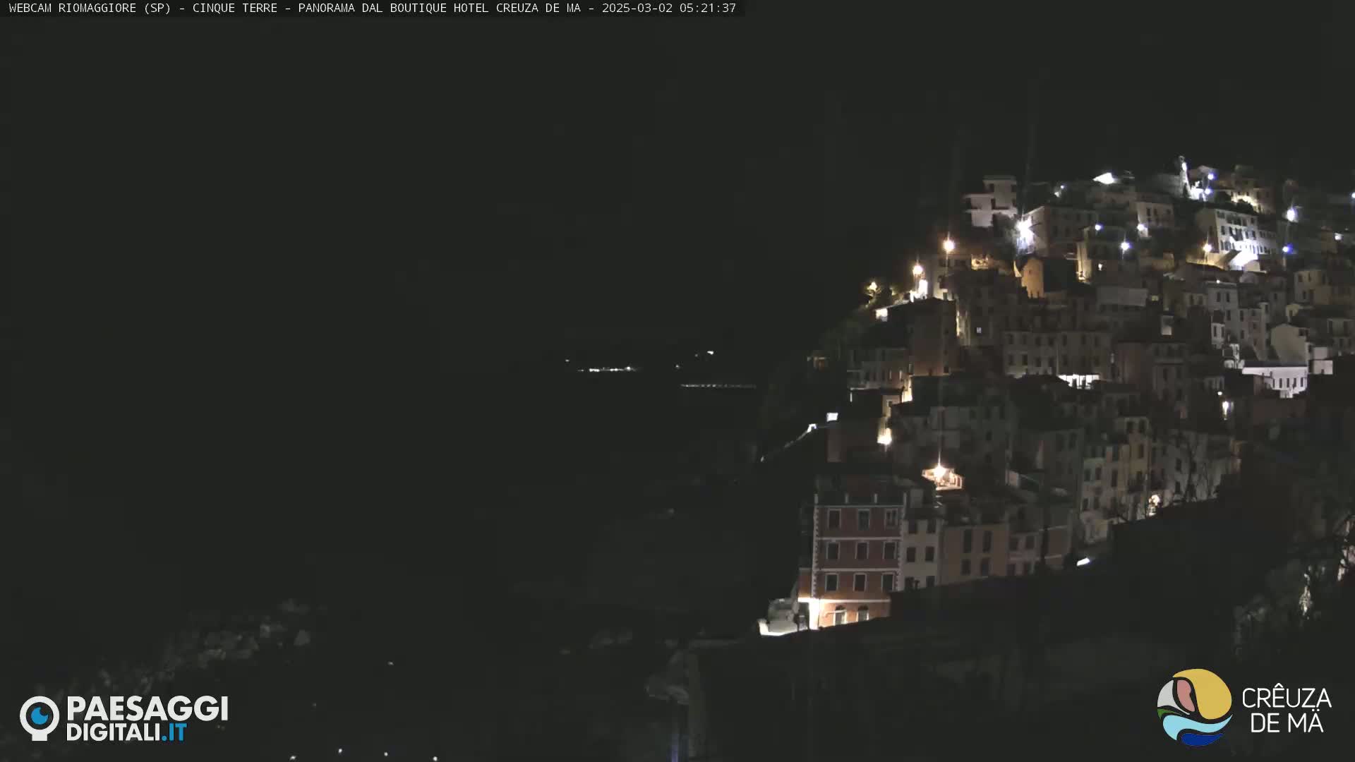 A hillside town is illuminated at night, overlooking a dark body of water under a clear, dark sky.