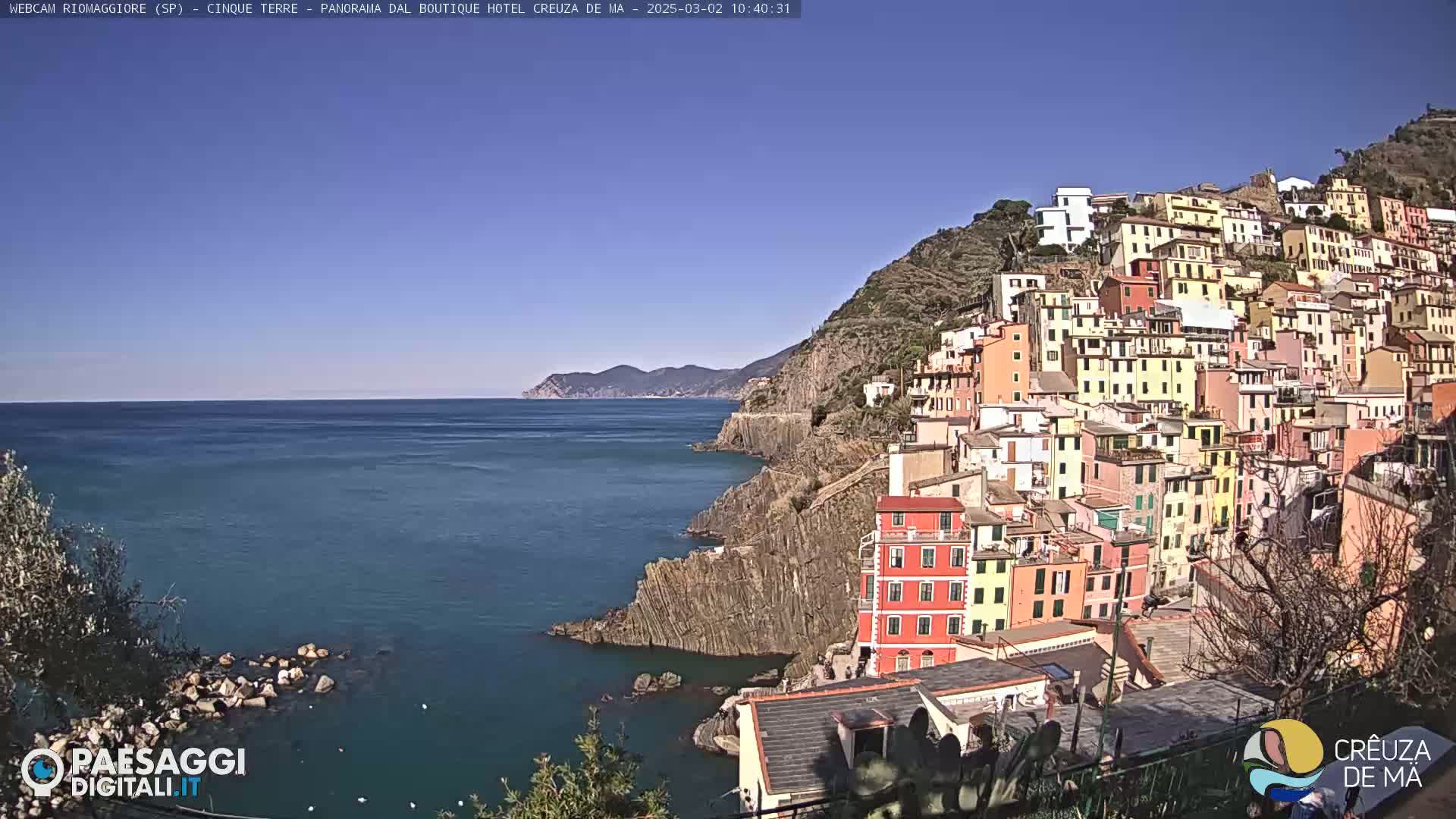 A colorful hillside village clings to a rocky coastline overlooking a calm blue sea under a clear blue sky.