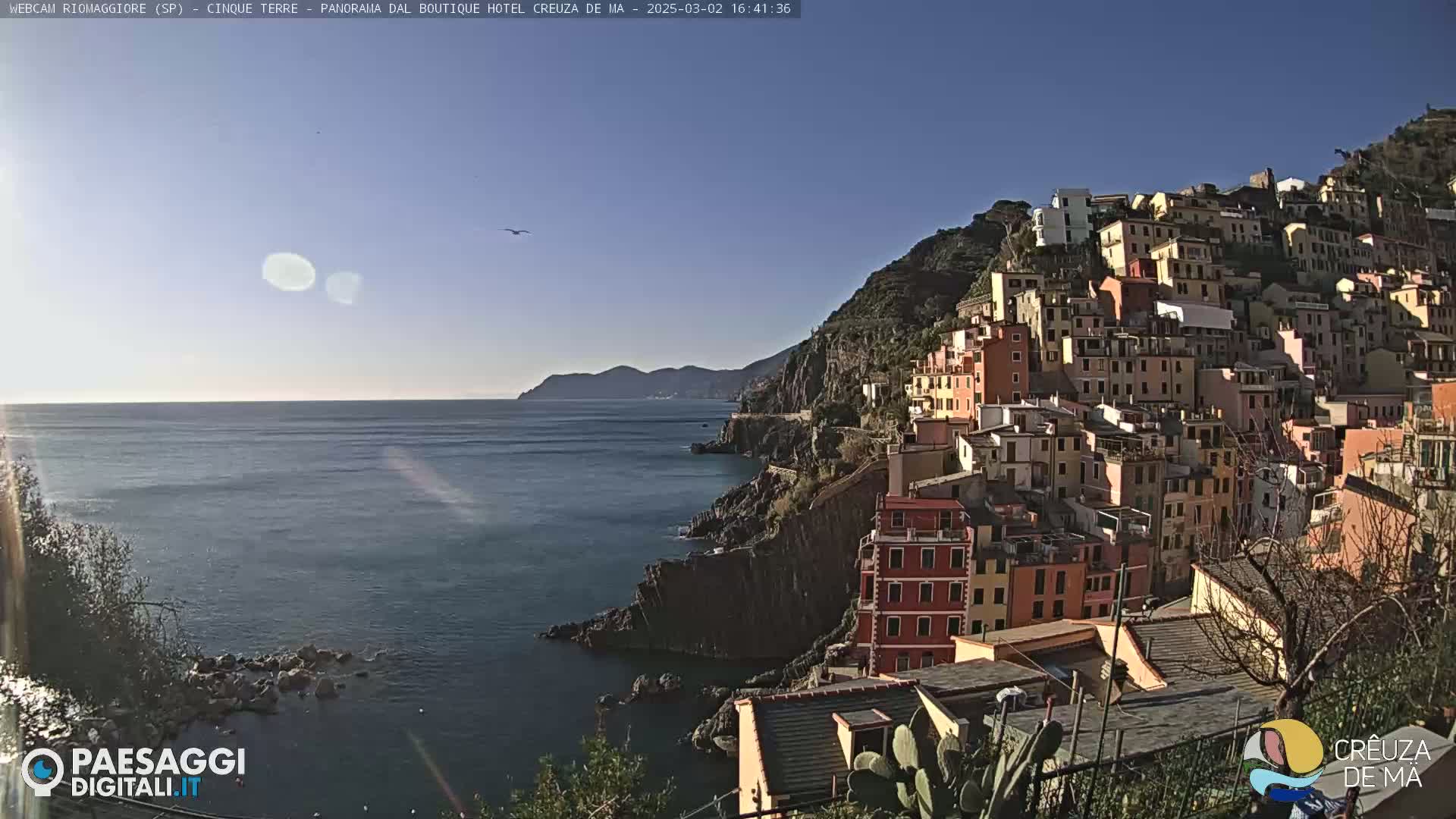 A colorful hillside town clings to a rocky coastline overlooking a calm, sunlit sea under a clear blue sky.