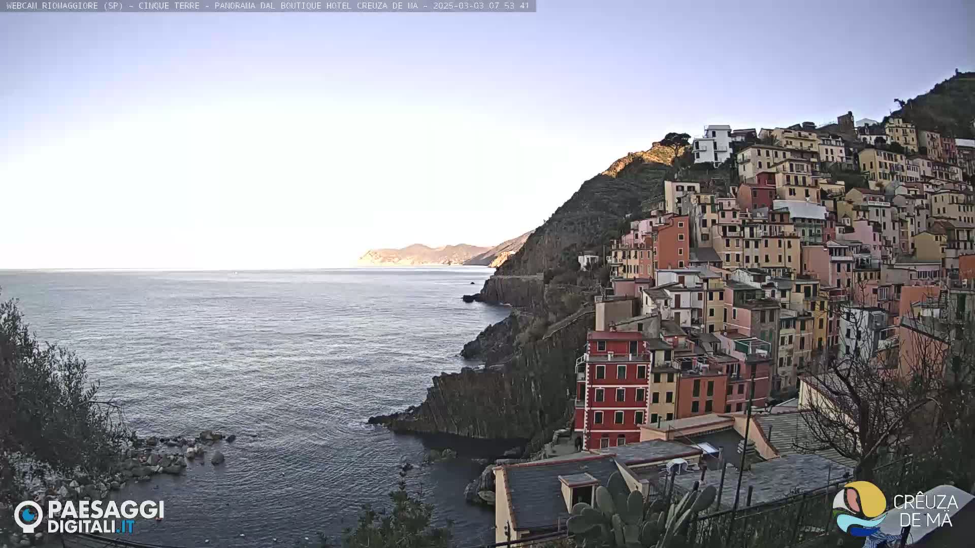 A colorful village clings to a steep cliffside overlooking a calm sea under a clear, bright sky.