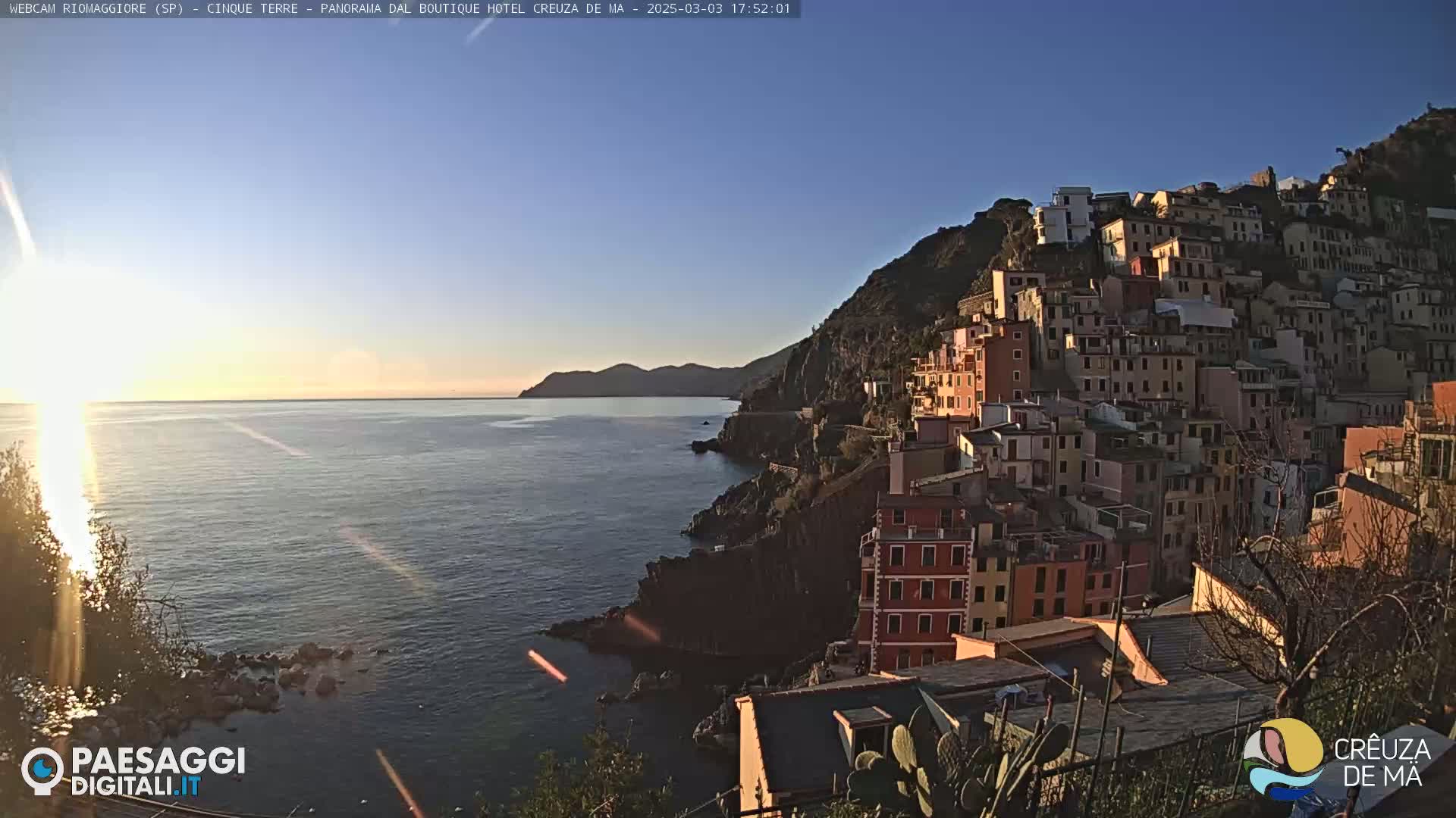 A coastal town of colorful buildings clings to a hillside overlooking a calm sea under a clear, bright sky.