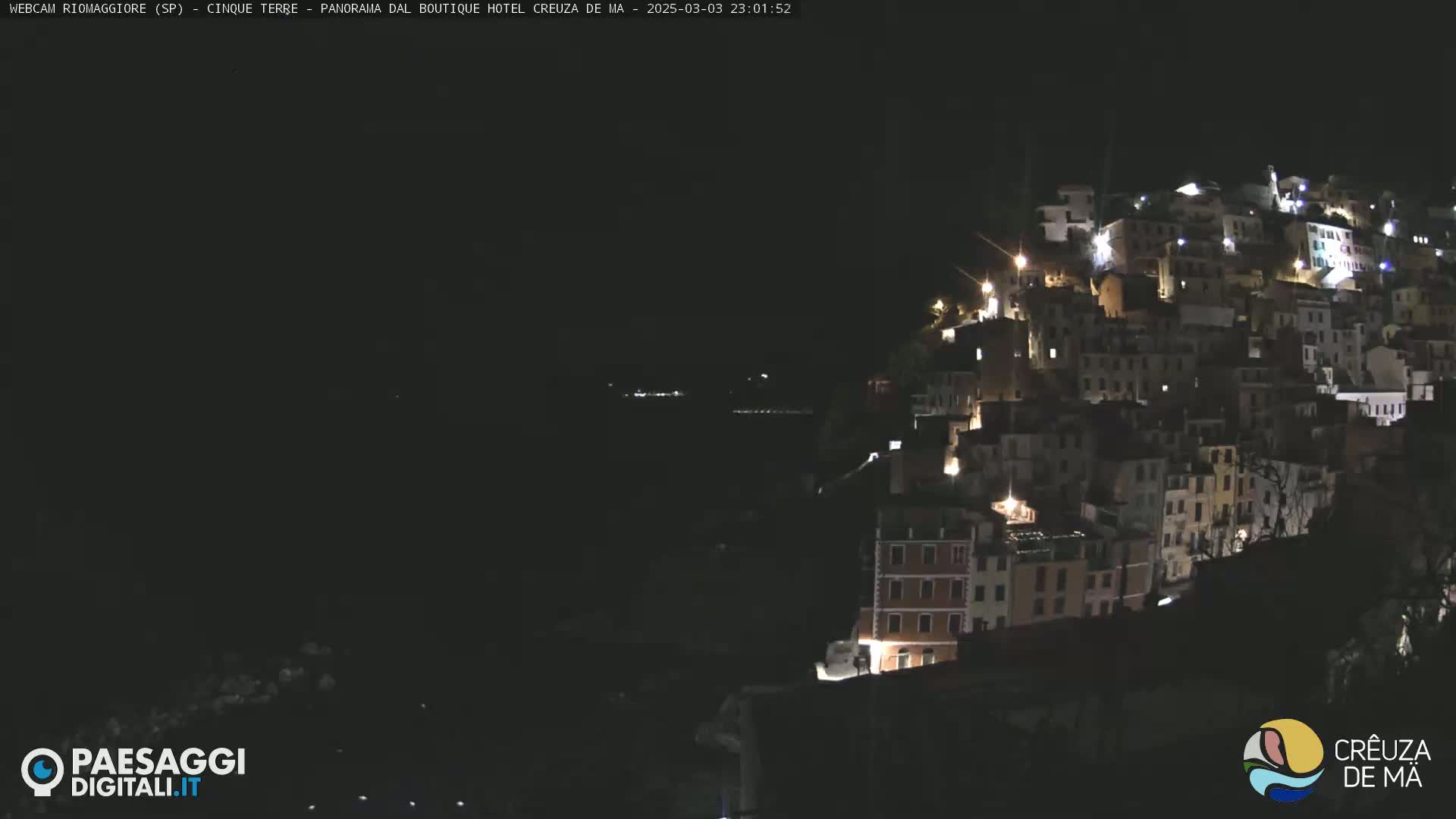 A hillside town is illuminated at night, overlooking a dark body of water under a clear, dark sky.