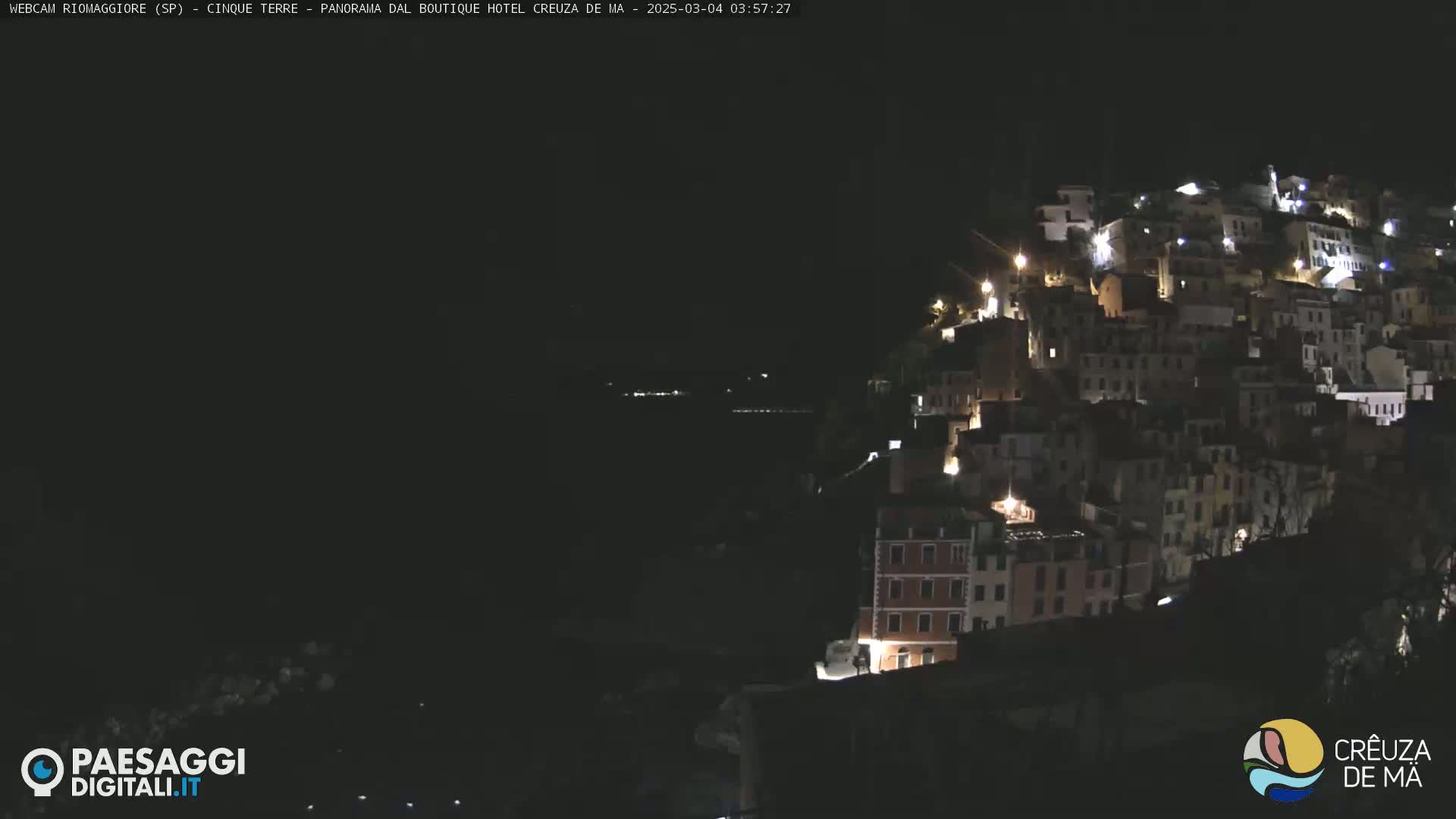 A nighttime, long shot of a hillside town illuminated by lights, overlooking a dark body of water.