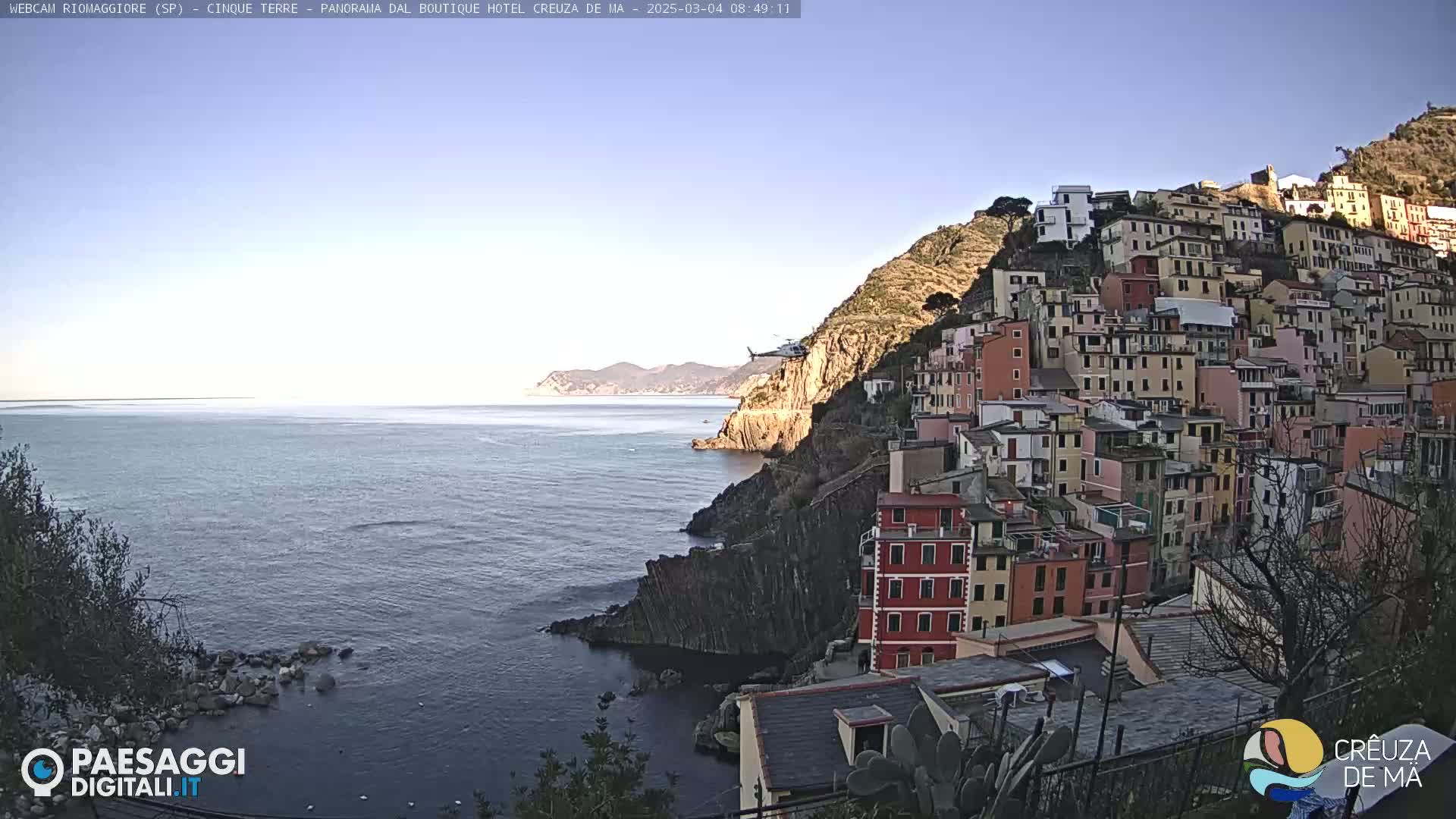 A helicopter flies over a clear, blue ocean next to a colorful hillside town built on a rocky cliff.