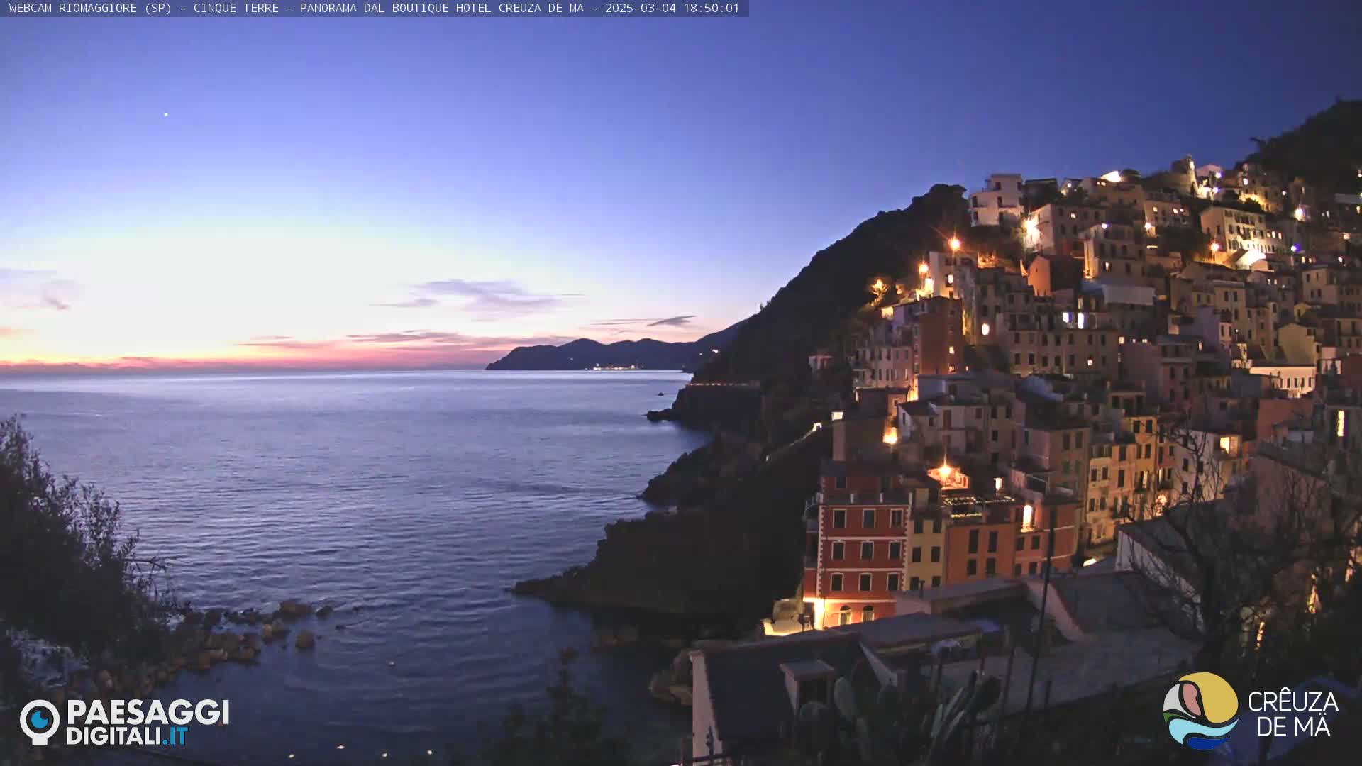 A twilight view shows a coastal town illuminated at night, nestled on a hillside overlooking a calm sea under a mostly clear sky.