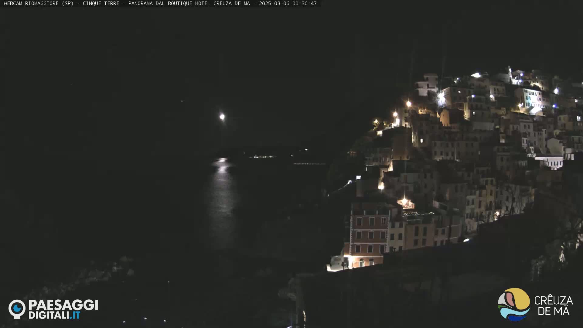 A nighttime view of a coastal town illuminated by lights, with a calm body of water reflecting the light of a moon in the sky.